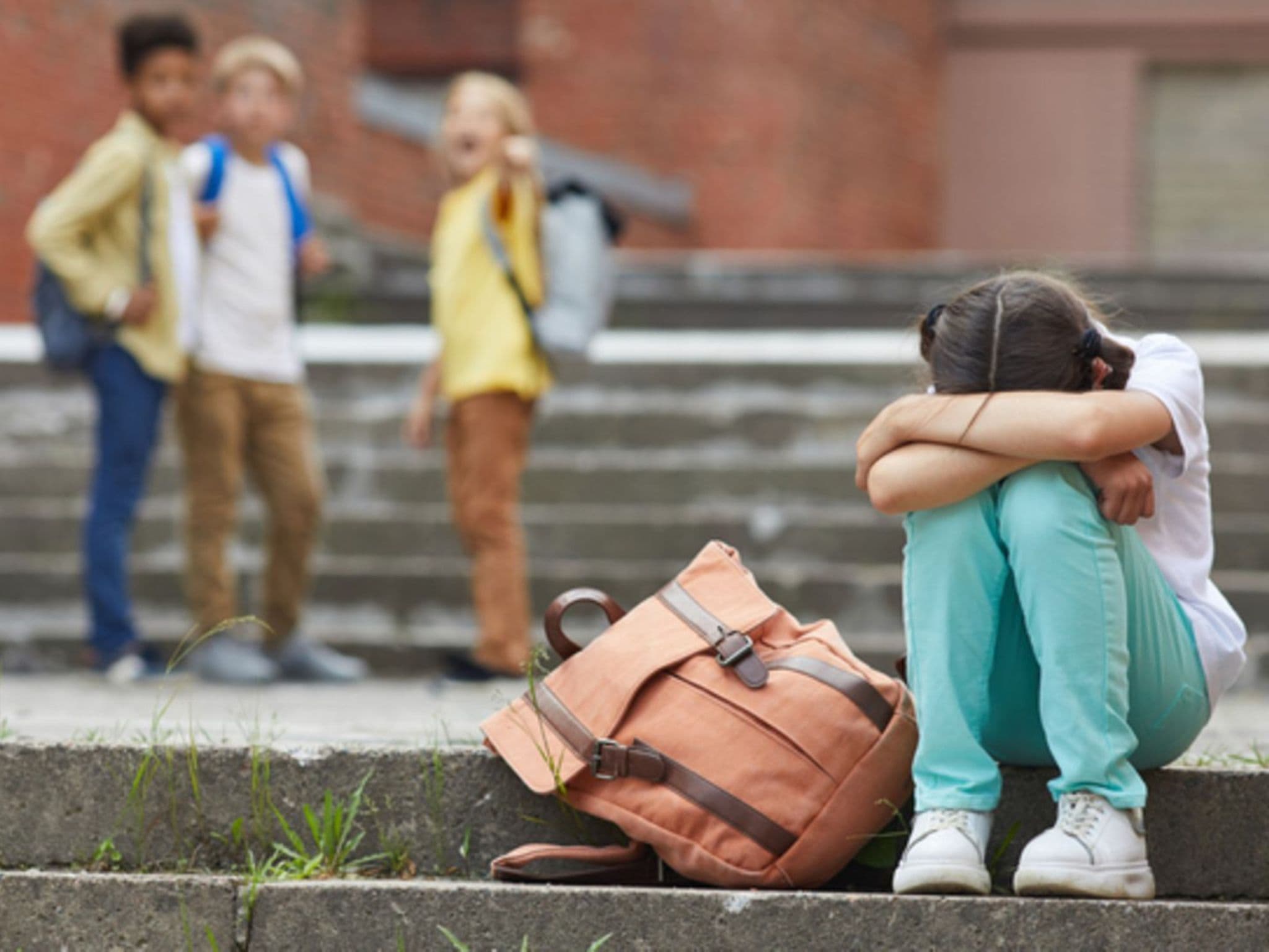 Mobbing: Ein Mädchen sitzt traurig auf dem Schulhof. Im Hintergrund sind mehrere Kinder, die auf sie zeigen.