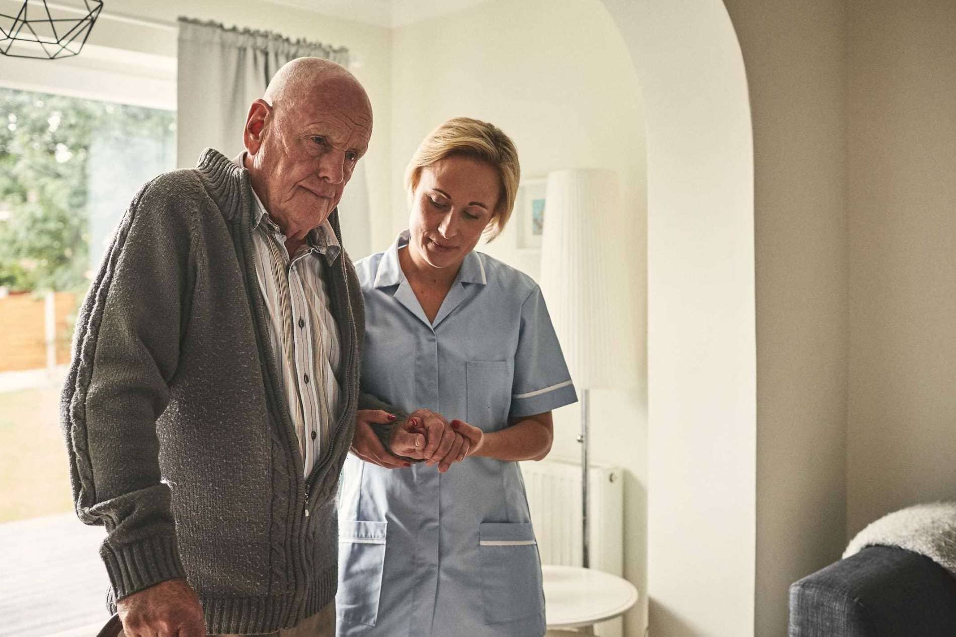 Häusliche Krankenpflege: Älterer Mann wird von einer Betreuerin am Arm geführt.