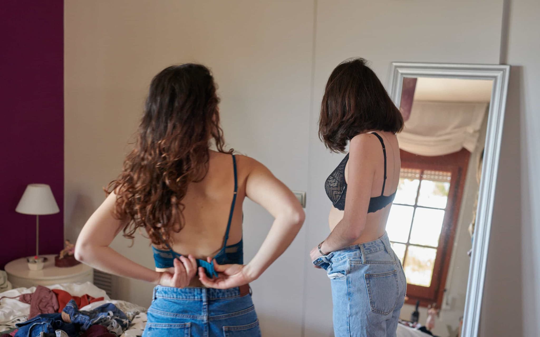 Magersucht: Zwei Frauen in Jeans und BH betrachten sich im Spiegel.