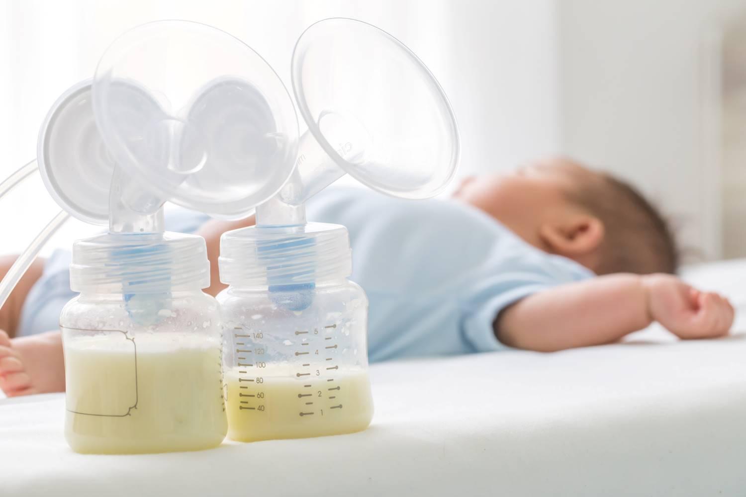 Milchpumpen: Zwei an eine Milchpumpe angeschlossene Fläschchen mit Muttermilch. Im Hintergrund liegt ein Baby.