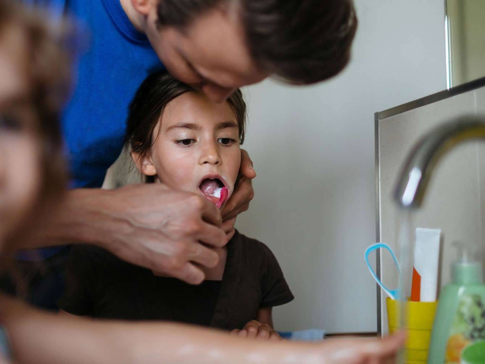 Zähneputzen: Vater putzt seiner Tochter am Waschbecken mit einer roten Zahnbürste die Zähne.