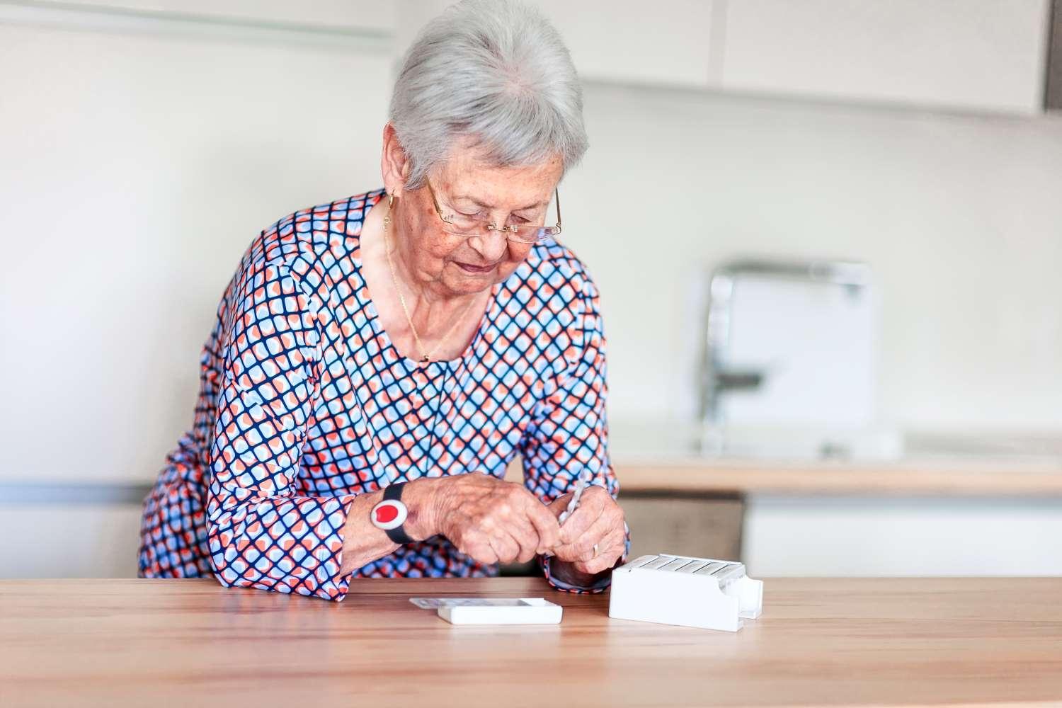 Hausnotruf: Ältere Frau füllt Tabletten in eine Schachtel und trägt ein Armband mit einem roten Notrufknopf am Handgelenk.