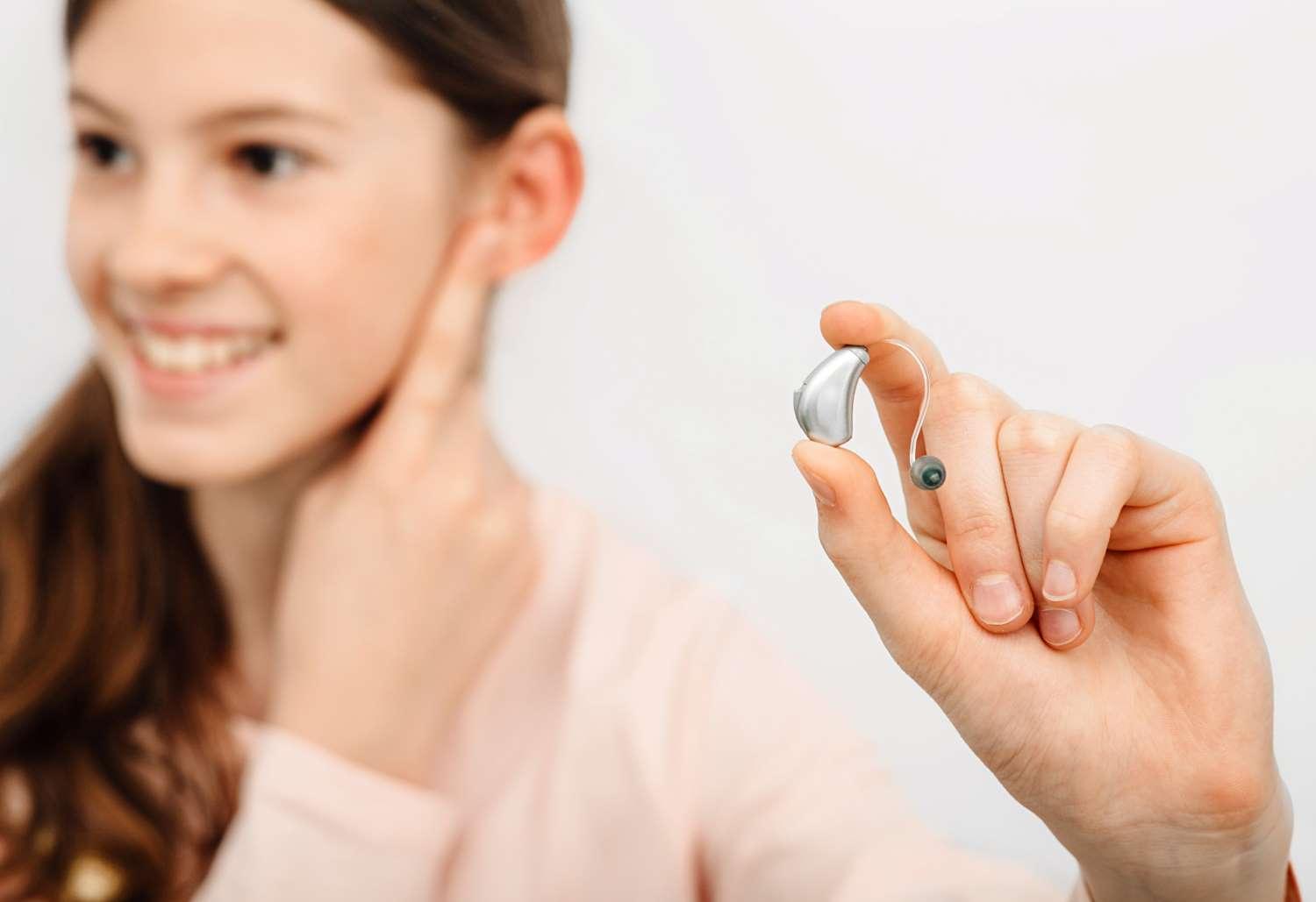 Hörgeräte: Junge Frau hält ein Hörgerät in ihrer Hand und fasst sich mit der anderen Hand ans Ohr.