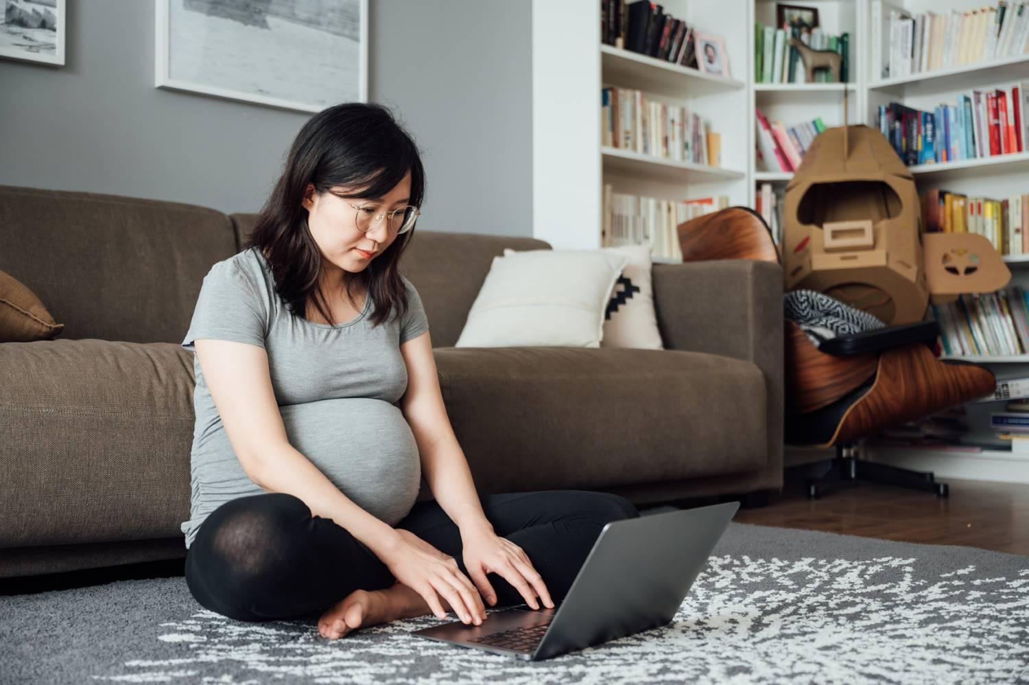 Mutterschutzfrist: Schwangere Frau sitzt in ihrem Wohnzimmer auf dem Boden und schaut auf ihr Notebook.
