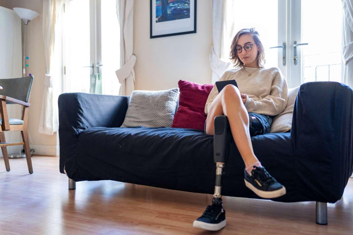 Prothesen: Junge Frau mit einer Beinprothese sitzt im Wohnzimmer auf einem Sofa und schaut auf ihr Smartphone.