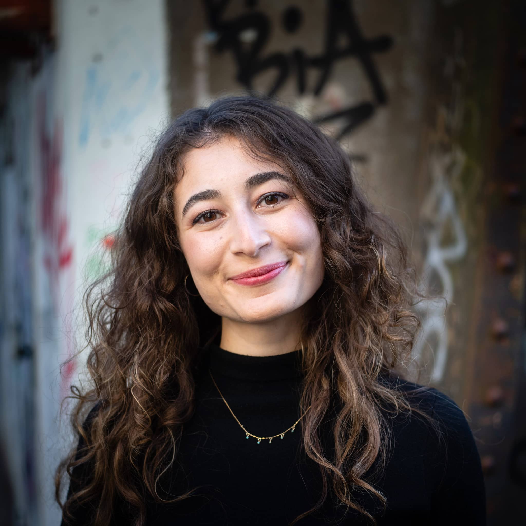Bild: Portrait Imke Anahita , eine Frau mit dunkelbraunen gelockten Haaren, Pulli schwarz, kleine Kette um