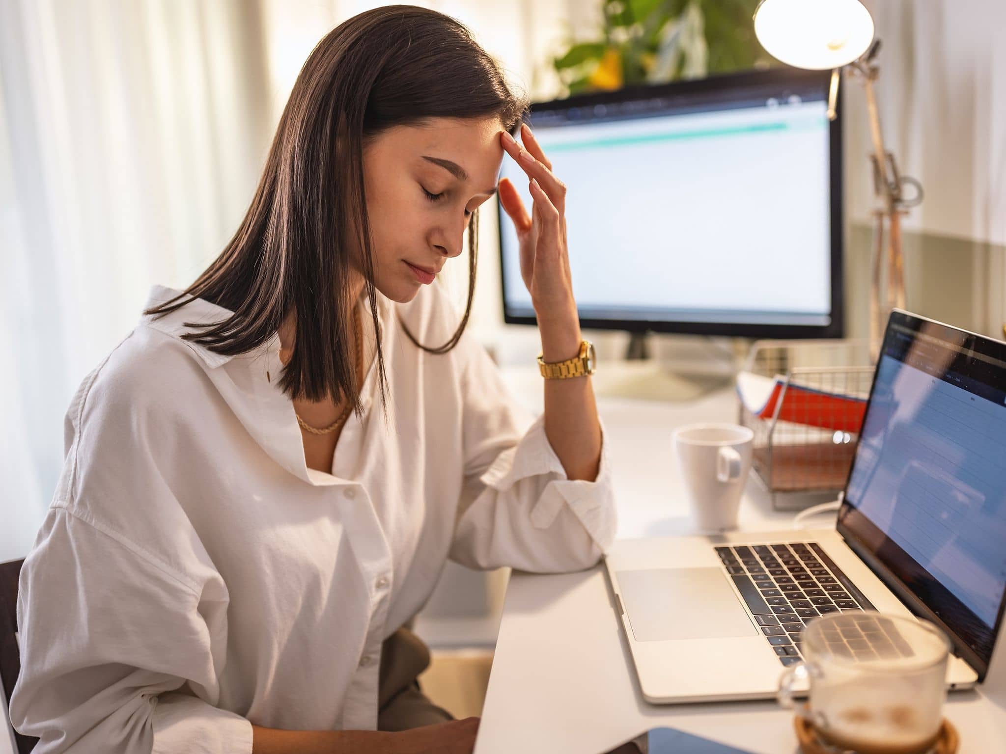 Person sitzt an einem Schreibtisch vor Laptop und Monitor und hält eine Hand an die Stirn.