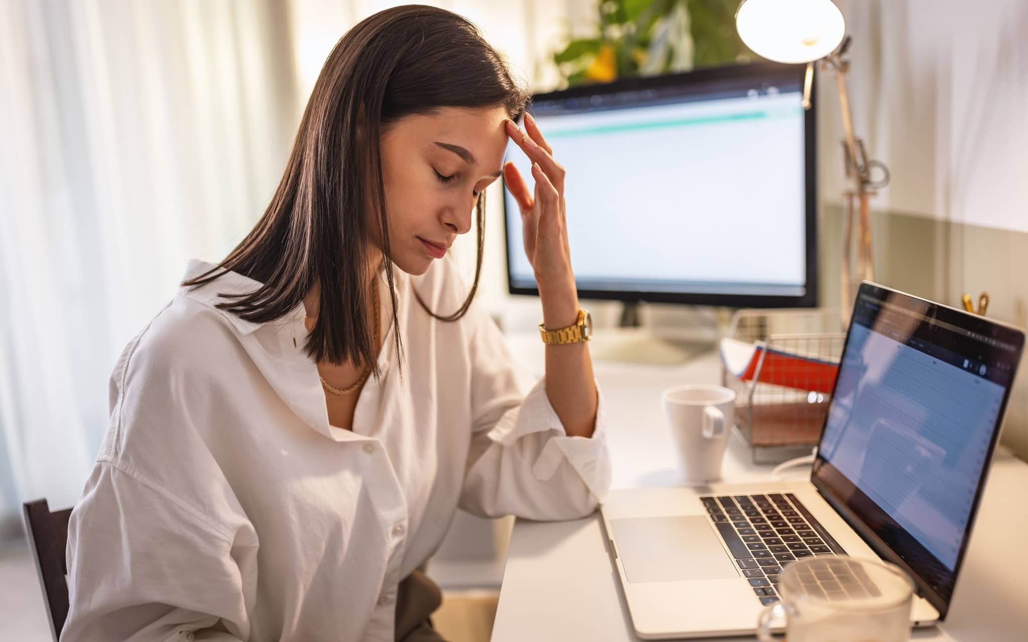 Person sitzt an einem Schreibtisch vor Laptop und Monitor und hält eine Hand an die Stirn.