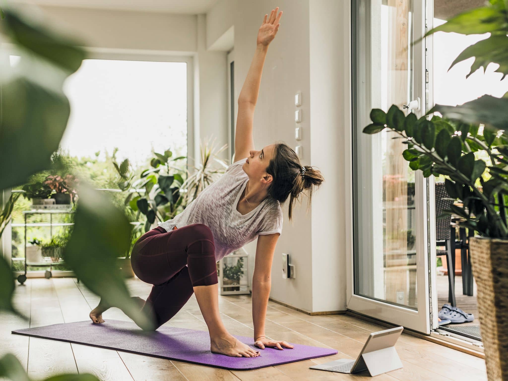 Frau, sportlich angezogen, Haare zusammengebunden macht im Wohnzimmer auf einer lila Sportmatte eine Yogaübung.