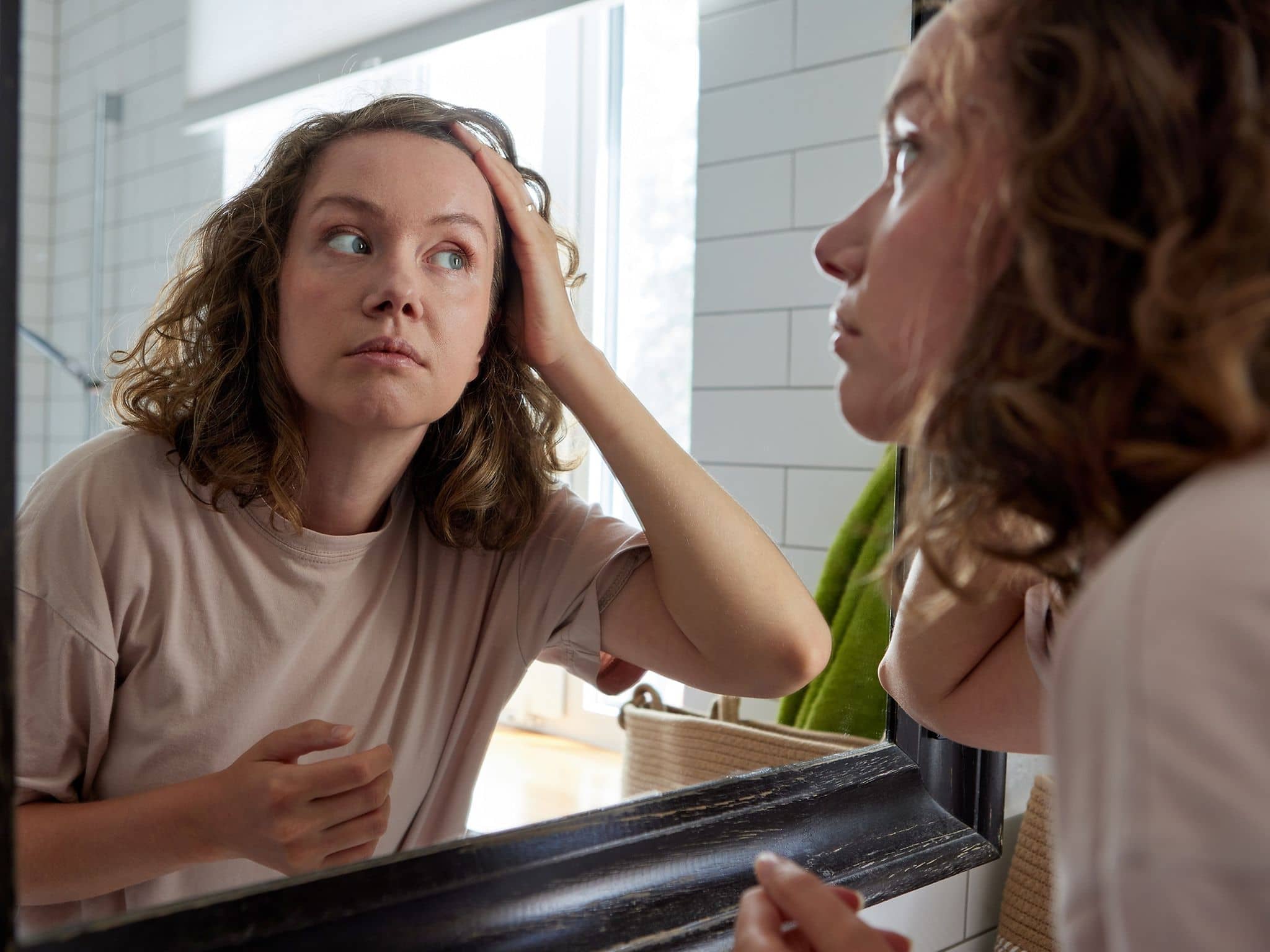 Person betrachtet ihre Haare im Badezimmerspiegel und streicht sich nachdenklich durch die Haare.