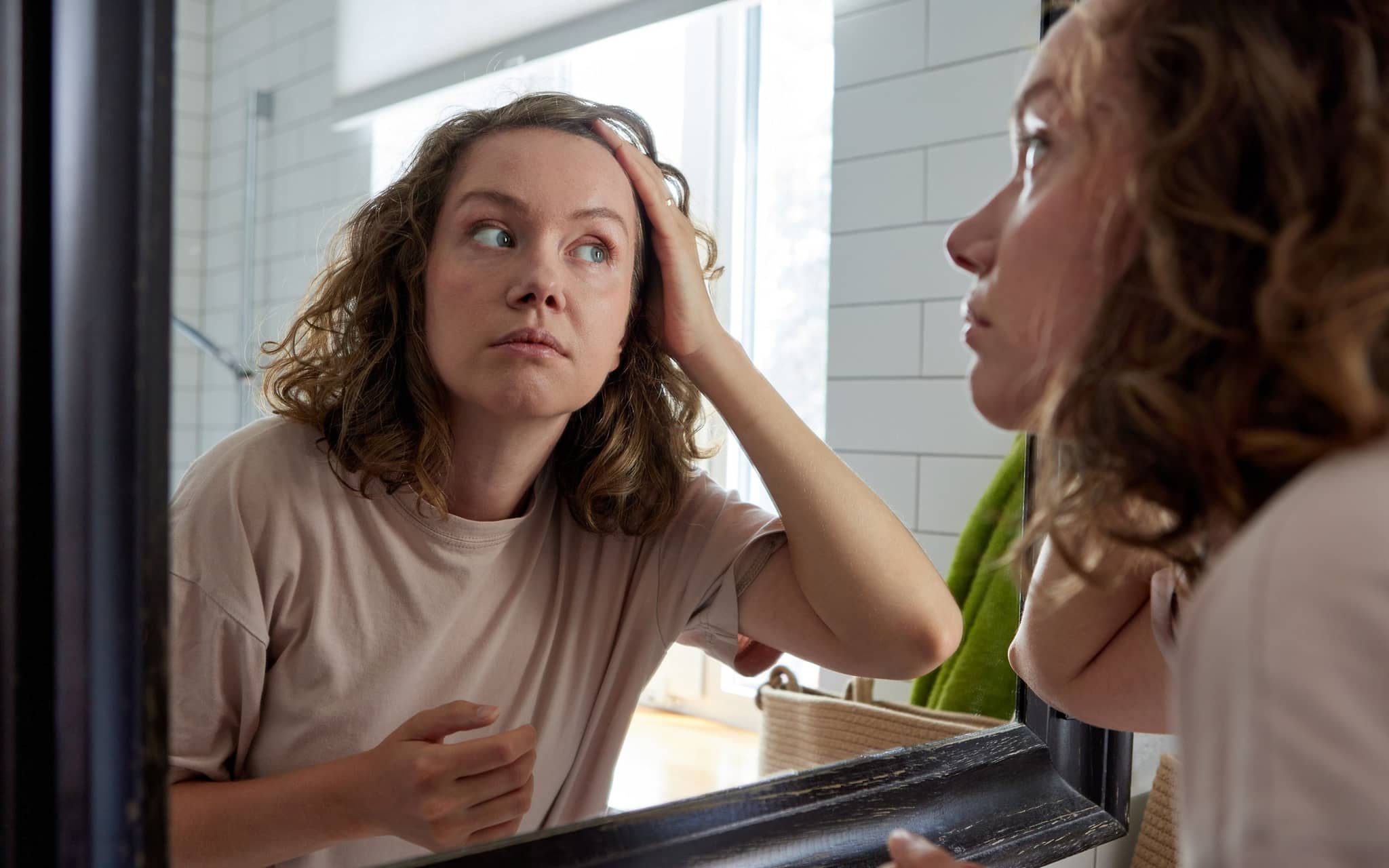 Person betrachtet ihre Haare im Badezimmerspiegel und streicht sich nachdenklich durch die Haare.