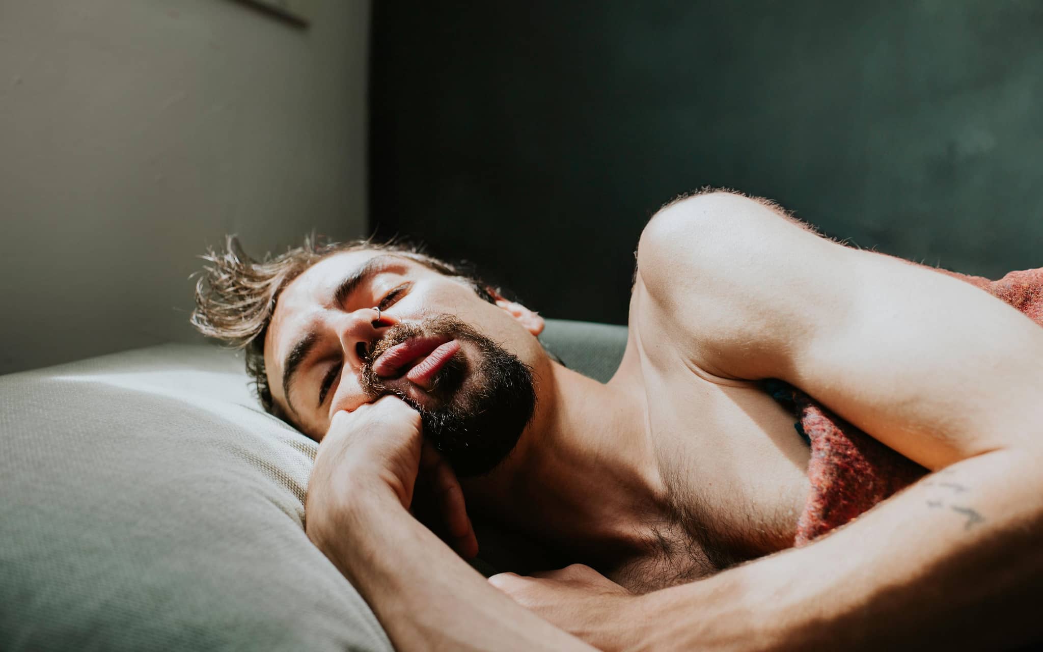 Mann schläft oberkörperfrei, halb zugedeckt und mit offenem Mund im Bett.