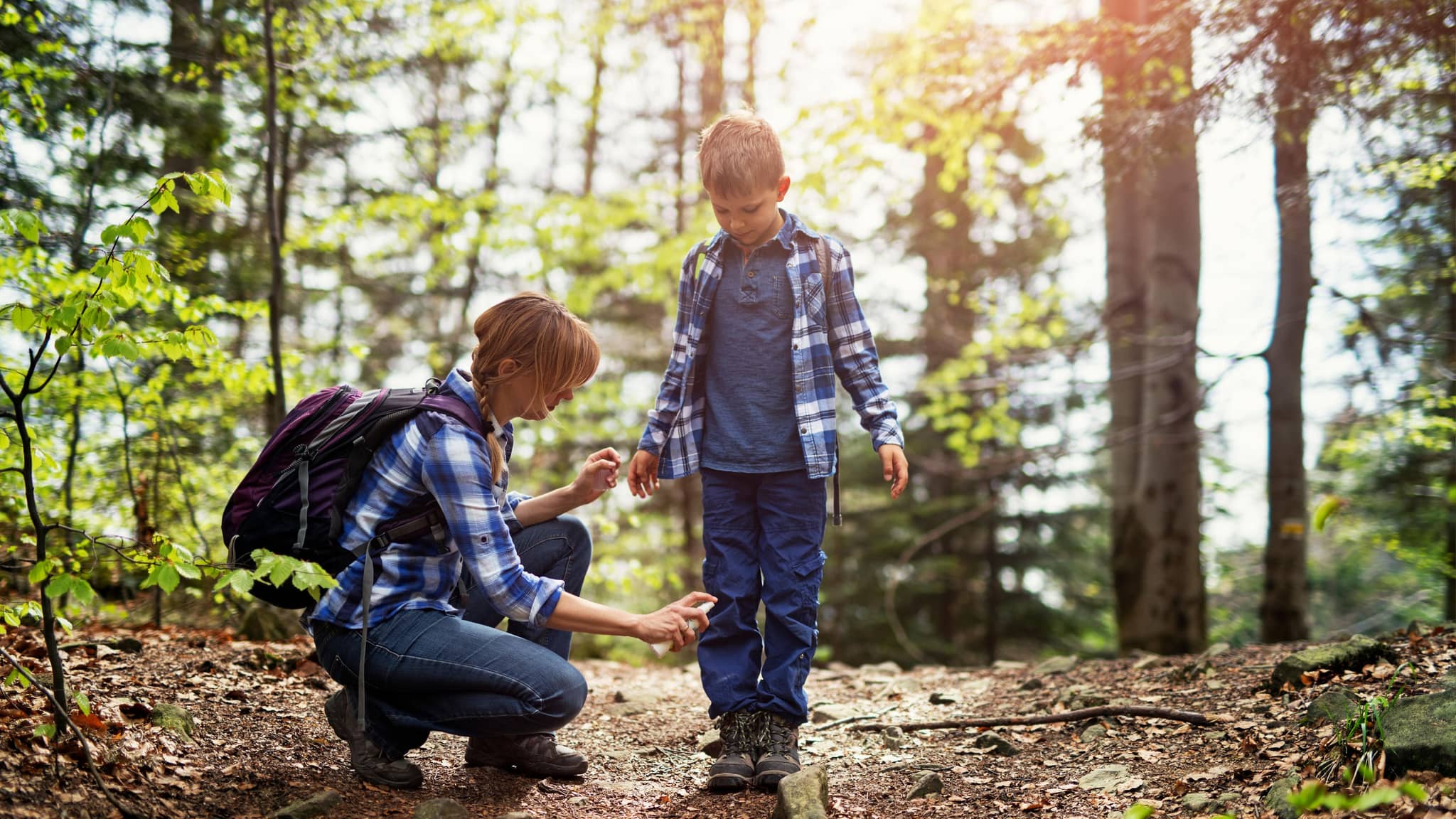 Mutter und Sohn wandern im Wald. Mutter trägt Zeckenschutzmittel auf die Beine des Sohnes auf.