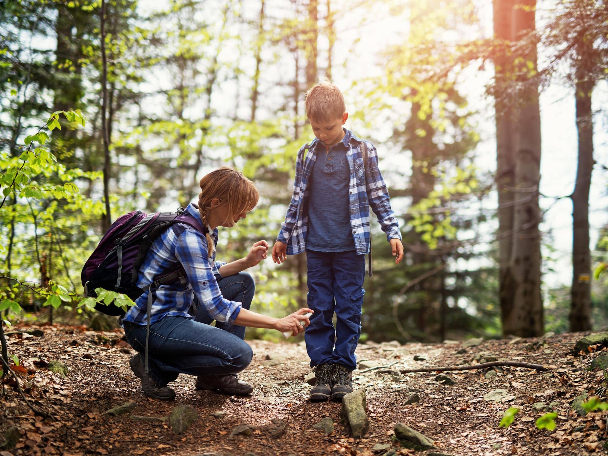 Mutter und Sohn wandern im Wald. Mutter trägt Zeckenschutzmittel auf die Beine des Sohnes auf.