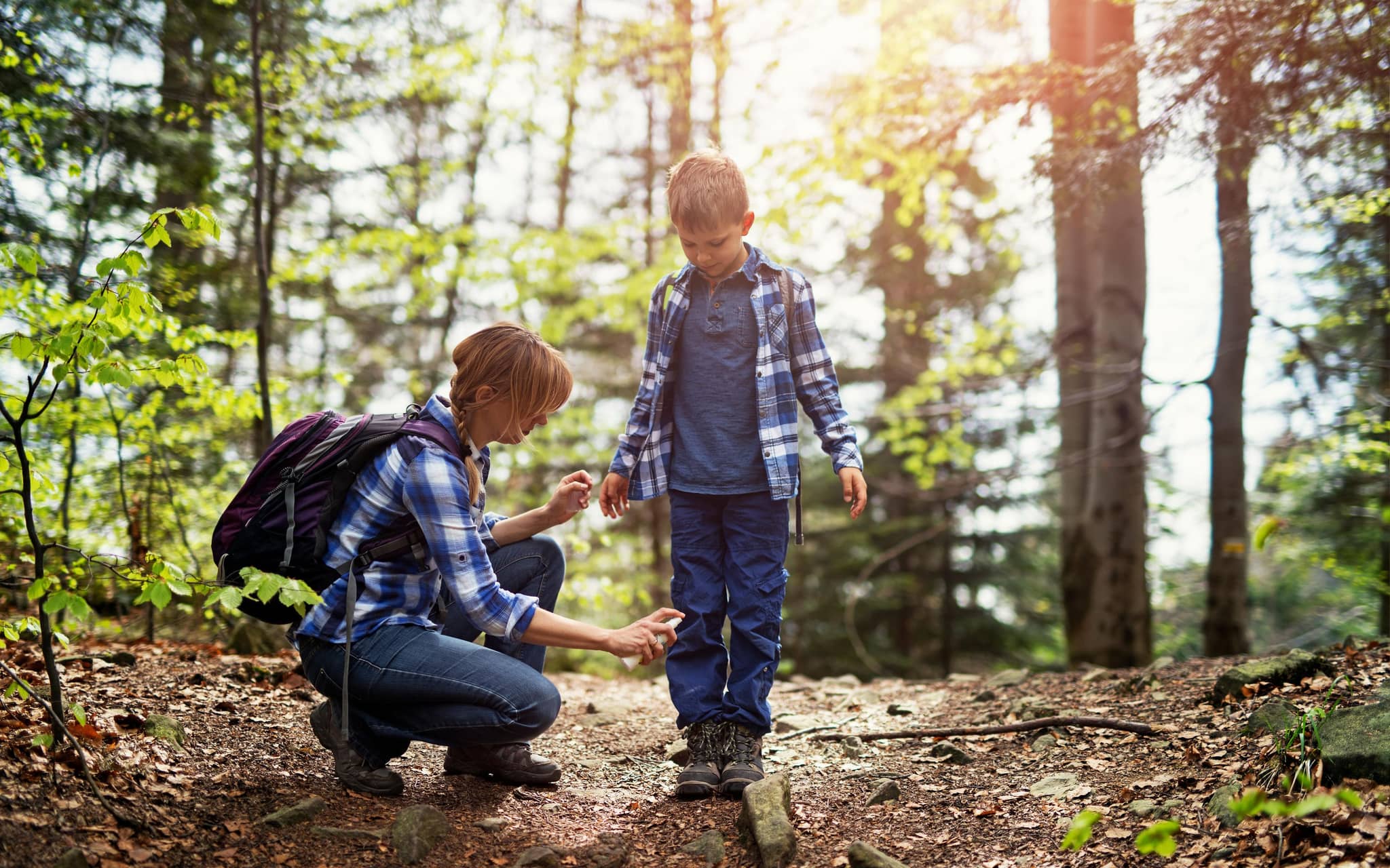 Mutter und Sohn wandern im Wald. Mutter trägt Zeckenschutzmittel auf die Beine des Sohnes auf.