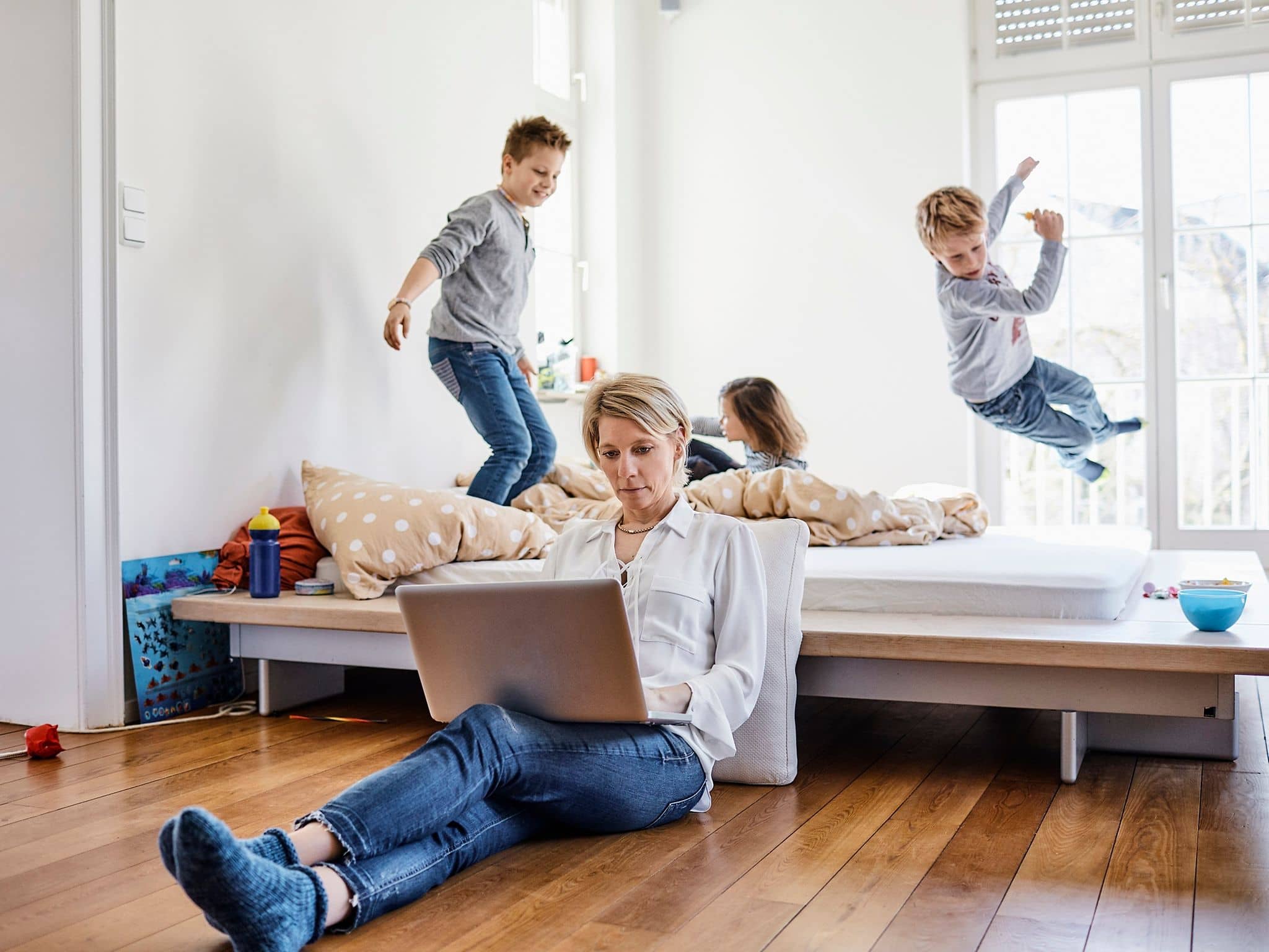 Frau arbeitet am Laptop, während drei Kinder im Hintergrund auf einem Bett spielen.