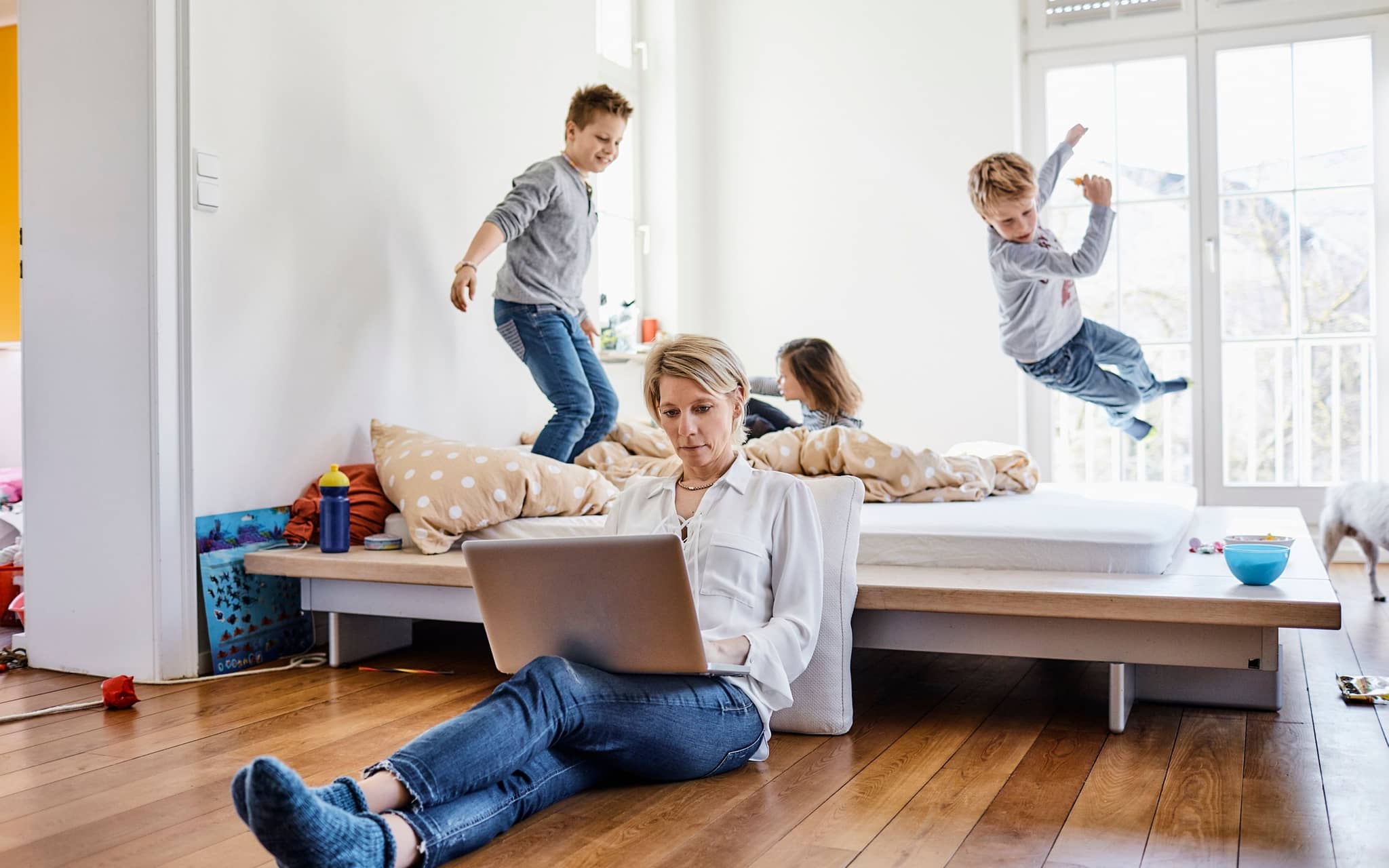 Frau arbeitet am Laptop, während drei Kinder im Hintergrund auf einem Bett spielen.