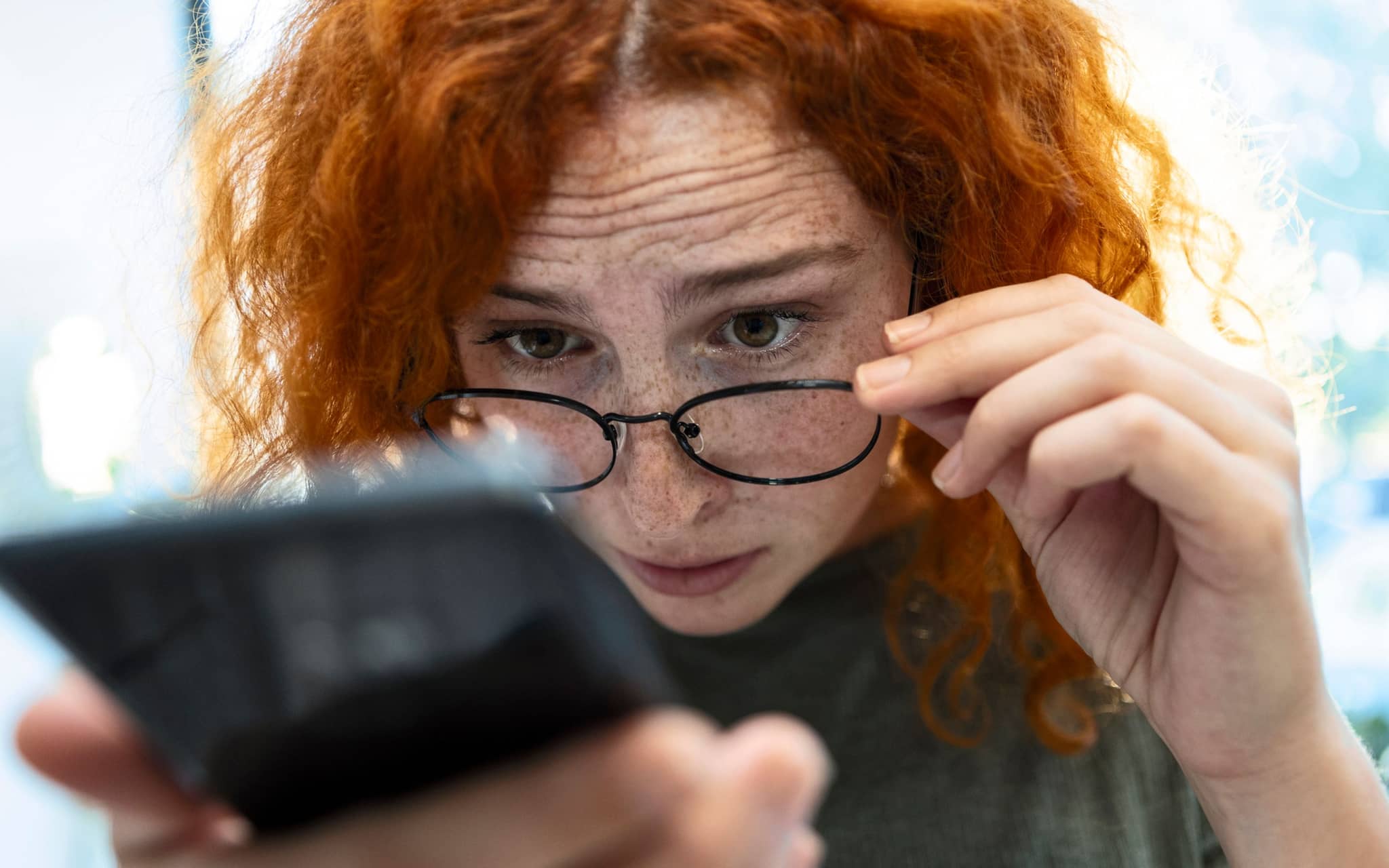 Frau schaut über ihre Brille auf ein Smartphone in ihrer Hand.