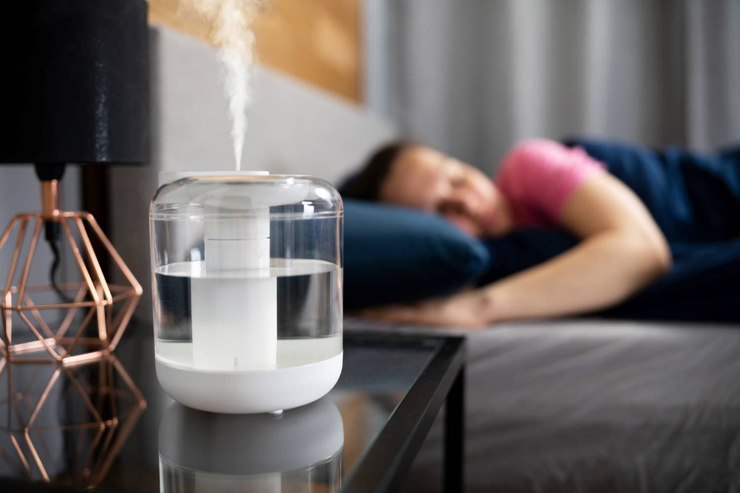 Raumluftbefeuchter: Auf dem Nachttisch steht ein aktiver Raumluftbefeuchter, im Hintergrund liegt eine Frau im Bett.