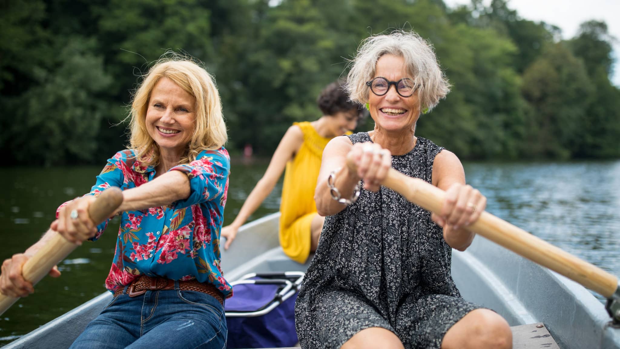 Zwei ältere Frauen rudern in einem Boot, mit einer dritten Frau im Hintergrund.
