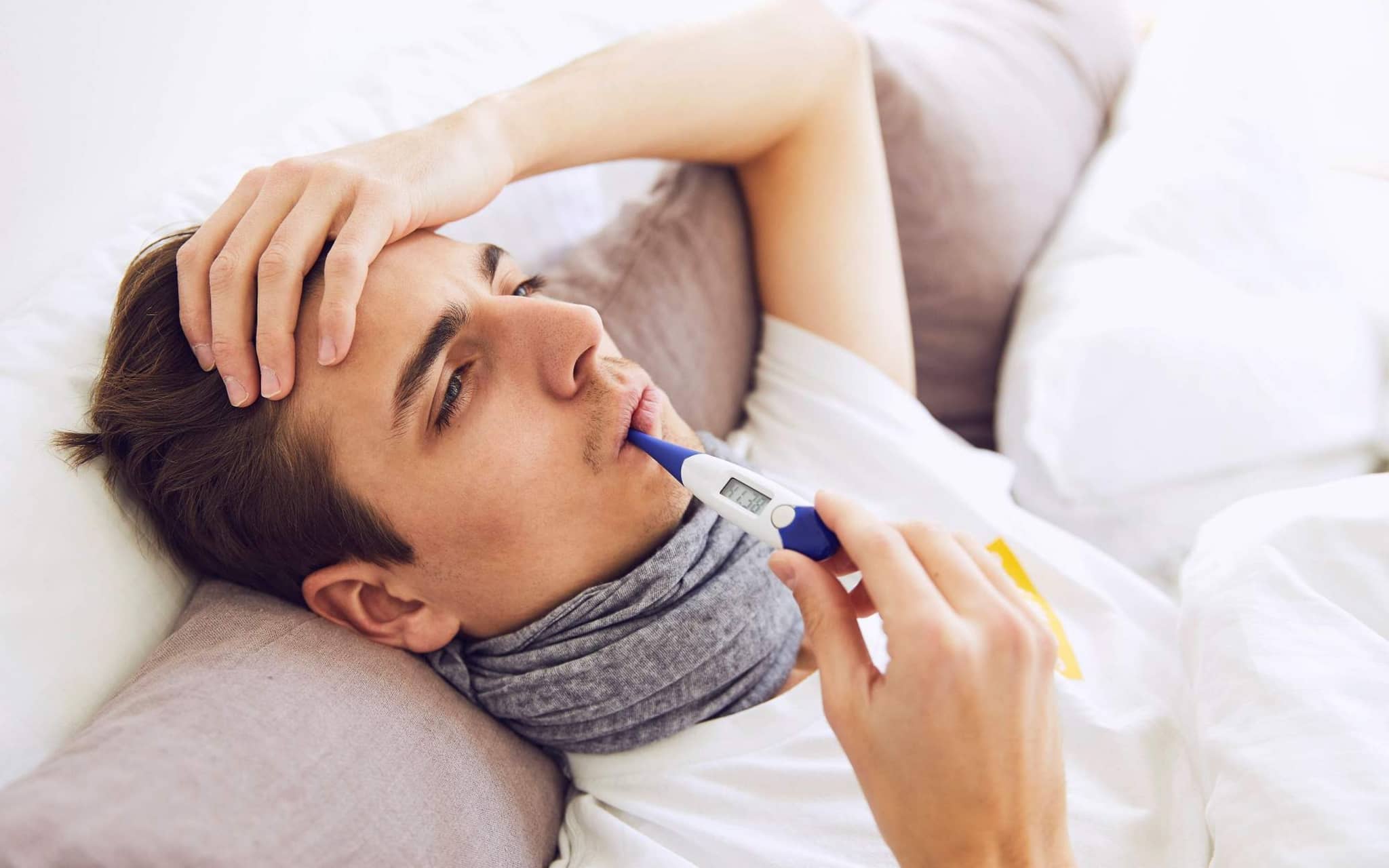 Lungenentzündung: Junger kranker Mann auf dem Sofa mit Halstuch hat Fieberthermometer im Mund und hält seine Hand an die Stirn.