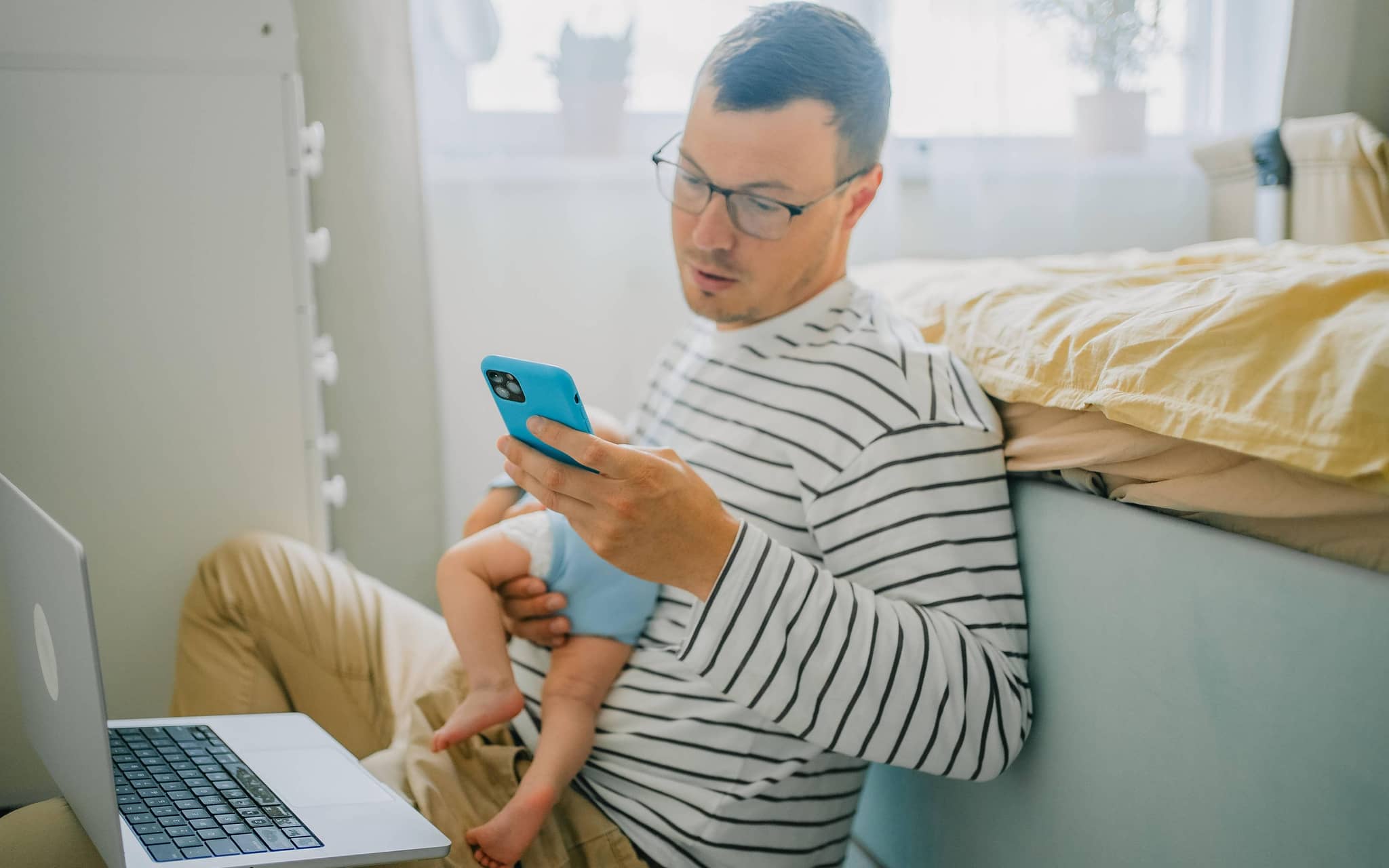 Ein beschäftigter Vater hält sein Baby auf dem Arm während er aufs Handy schaut und einen Laptop auf dem Schoß hat.