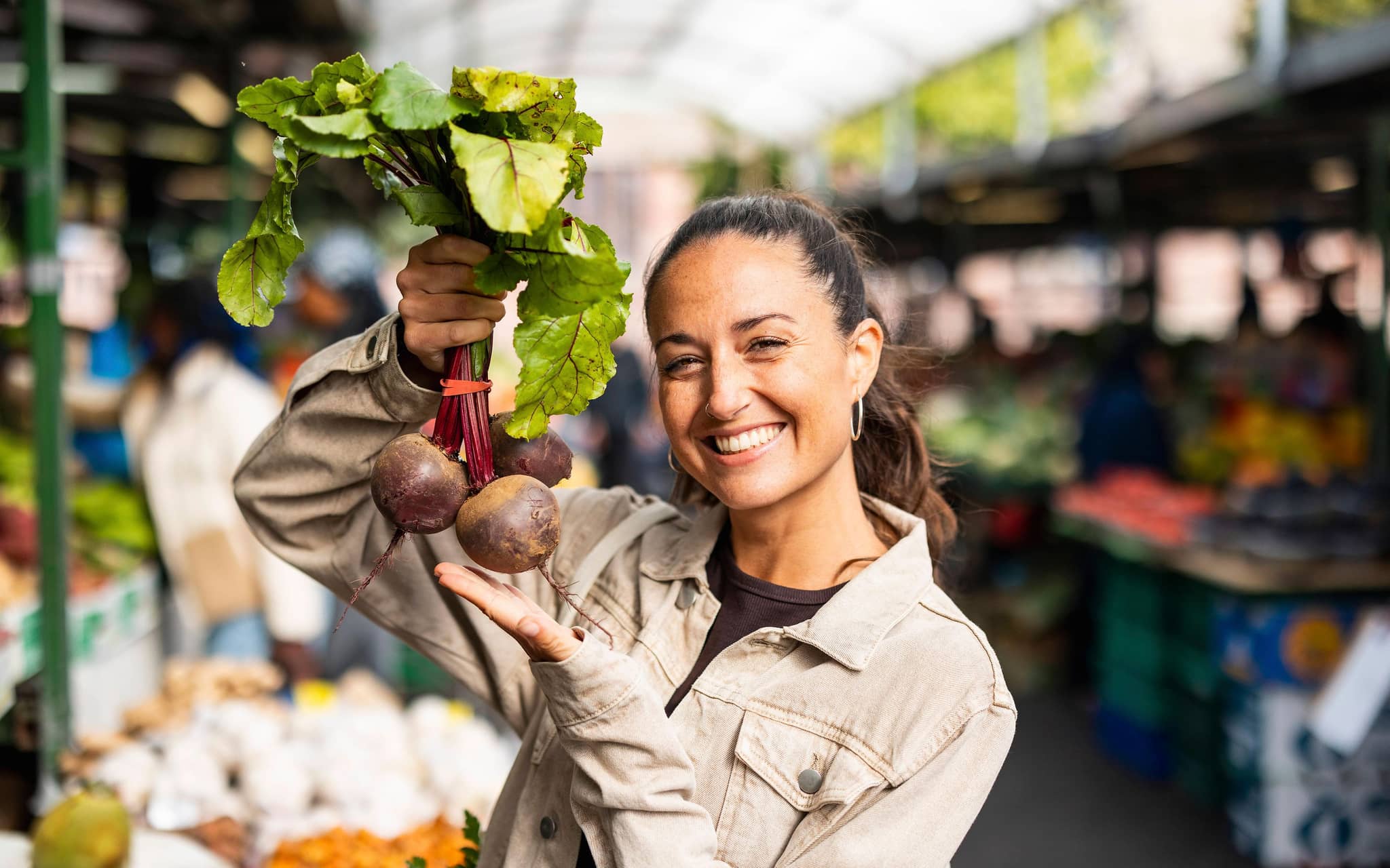 Frau hält lächelnd Rote Bete vor einem Marktstand in die Höhe.