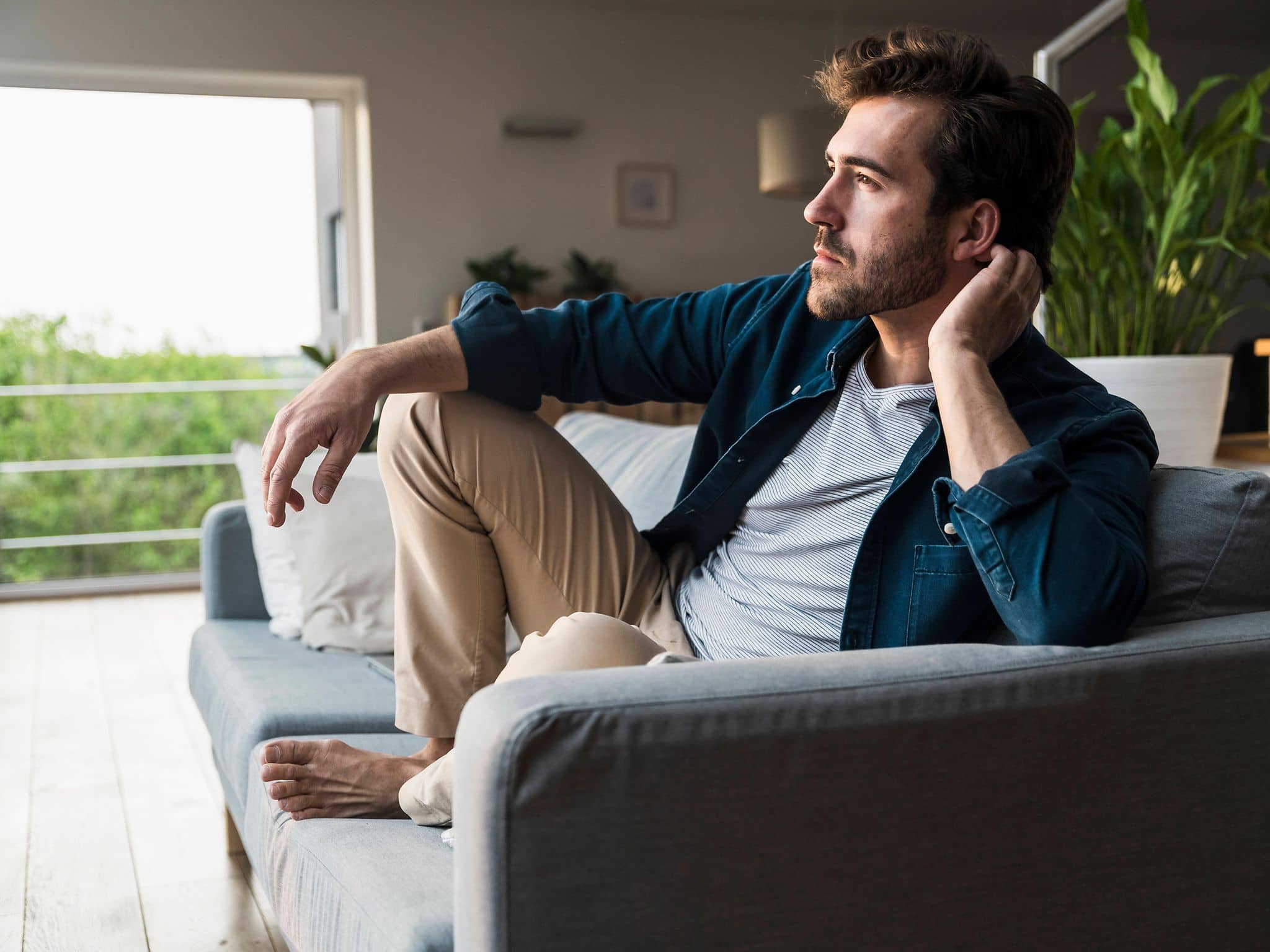 Mann im Jeanshemd sitzt mit angezogenen Beinen auf dem Sofa und guckt nachdenklich