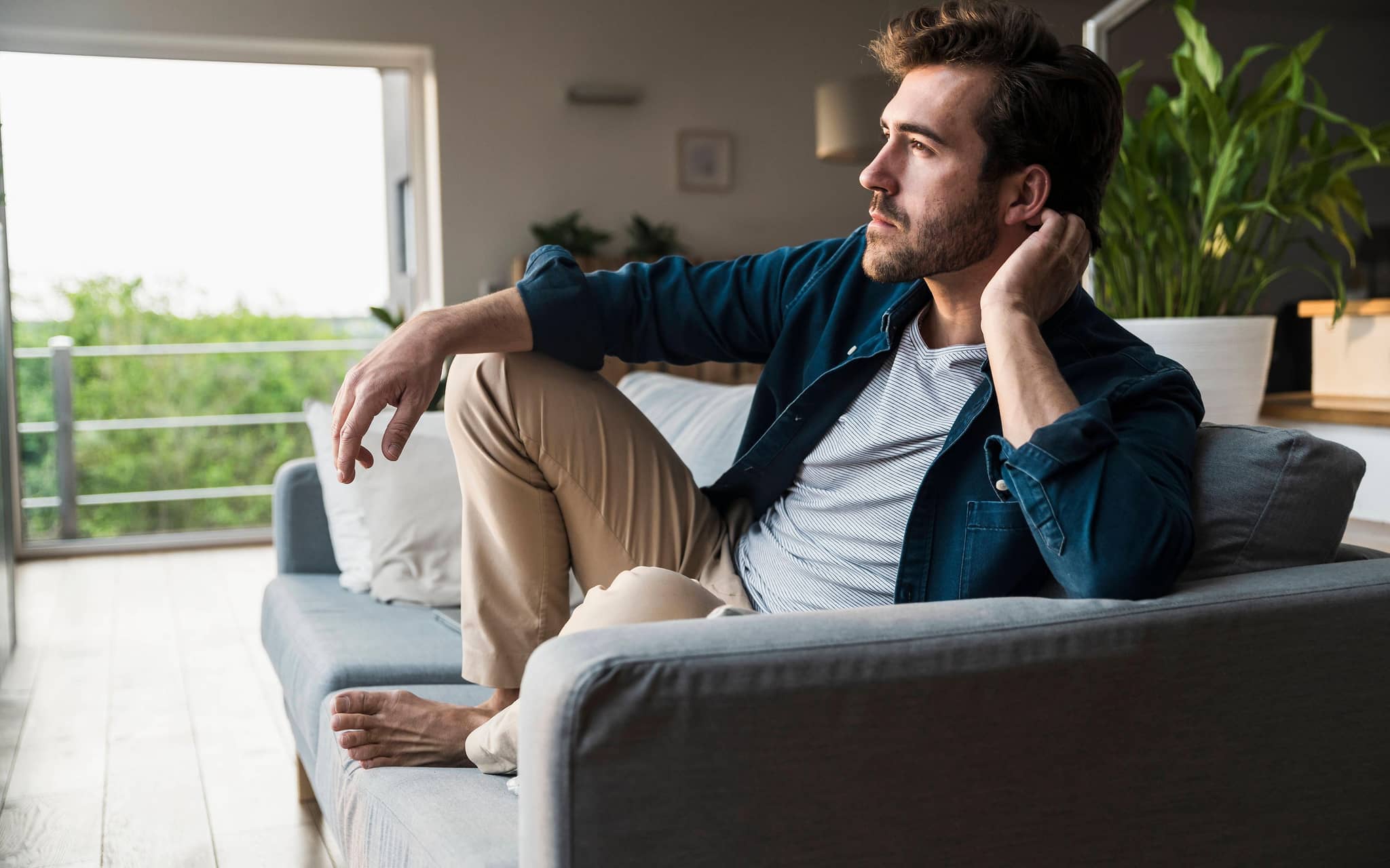 Mann im Jeanshemd sitzt mit angezogenen Beinen auf dem Sofa und guckt nachdenklich