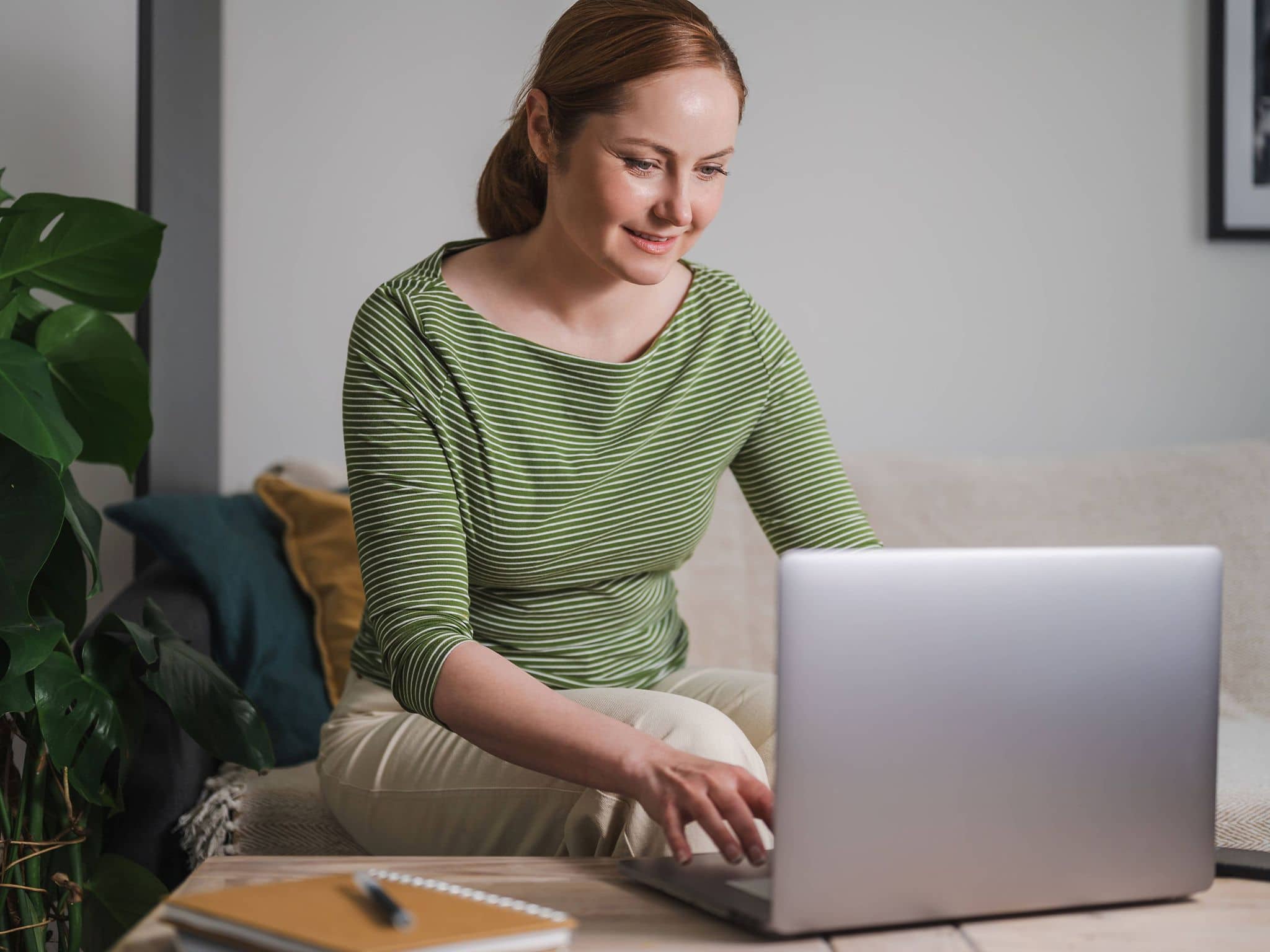Eine Frau mit gestreiftem Oberteil sitzt im Wohnzimmer und nutzt ihren Laptop.