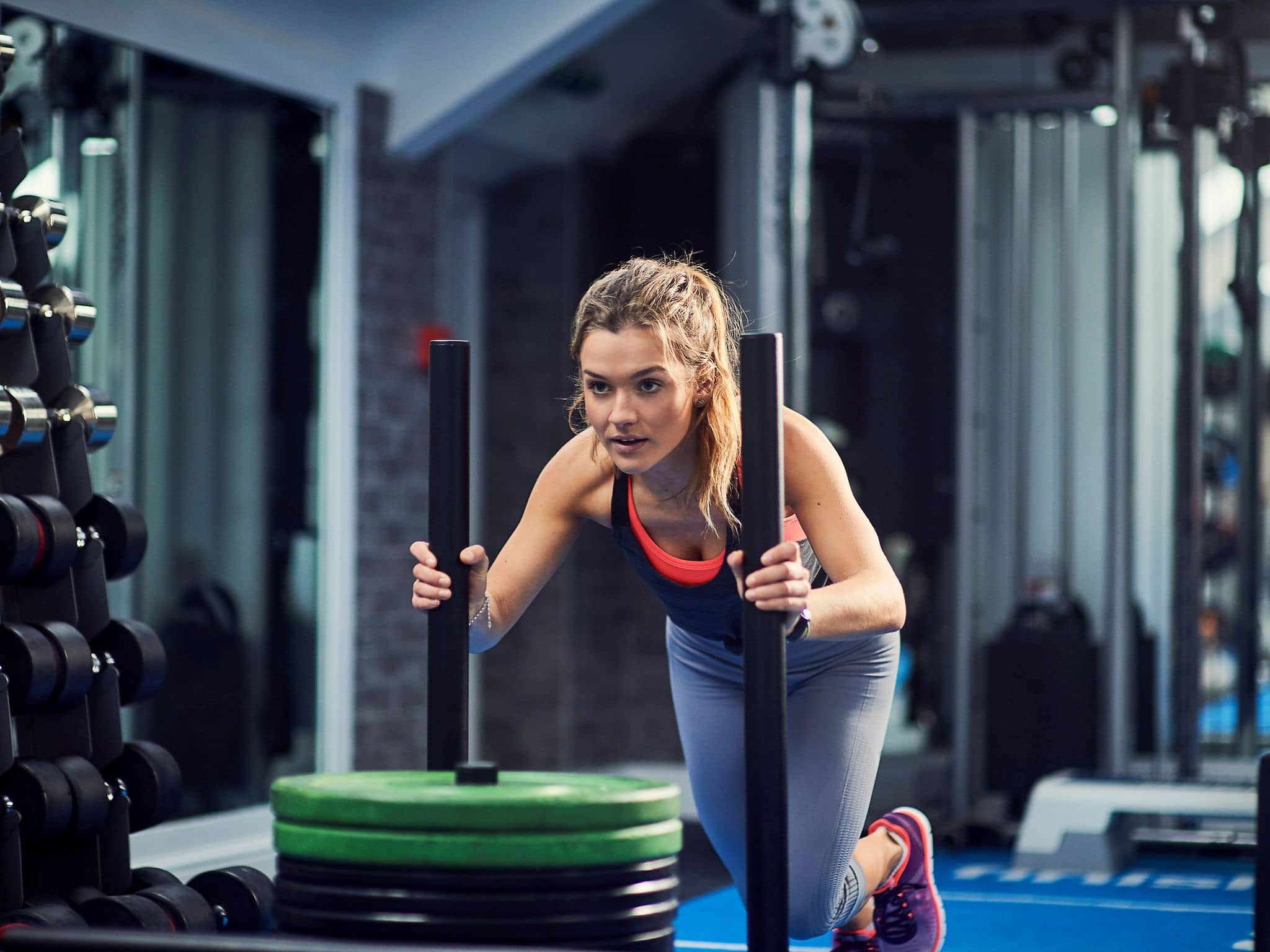 Eine Frau macht die Hyrox-Übung Sled-Push und schiebt einen Schlitten mit Gewichten in einem Fitnessstudio.