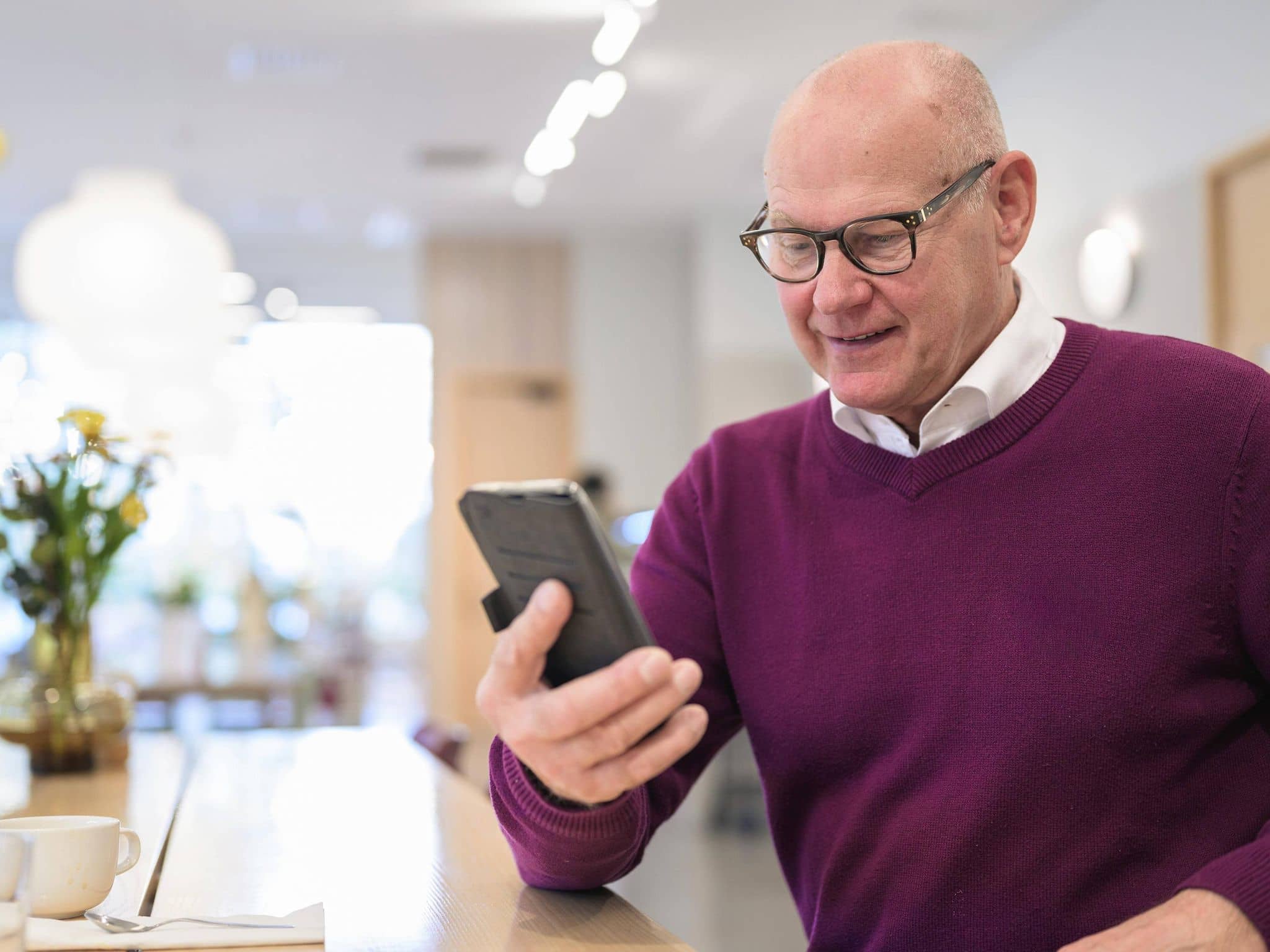 Ein älterer Mann mit rotem Pullover schaut in der Küche auf sein Handy.
