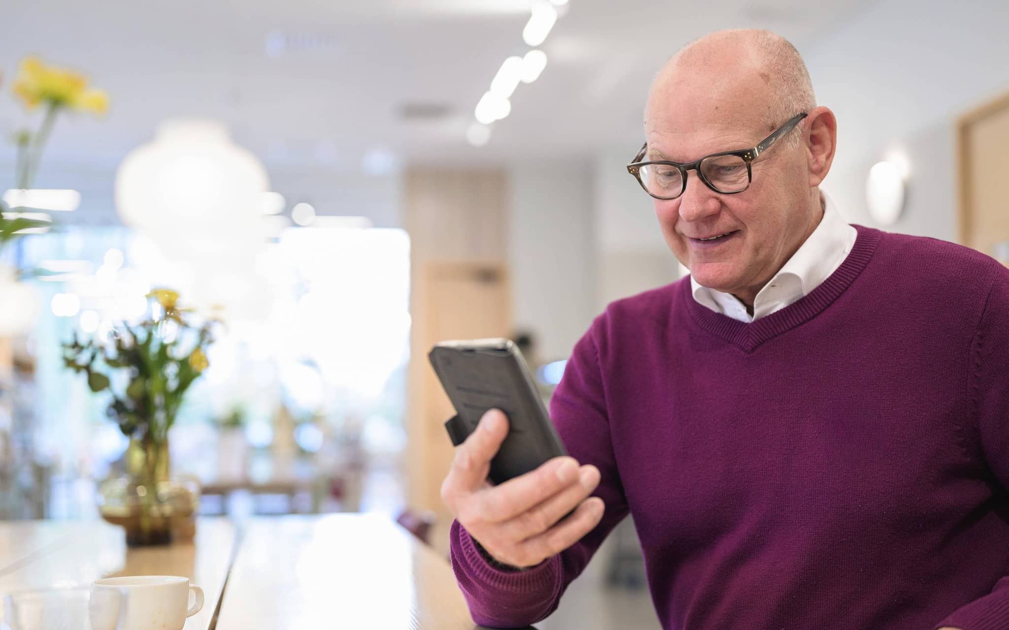 Ein älterer Mann mit rotem Pullover schaut in der Küche auf sein Handy.