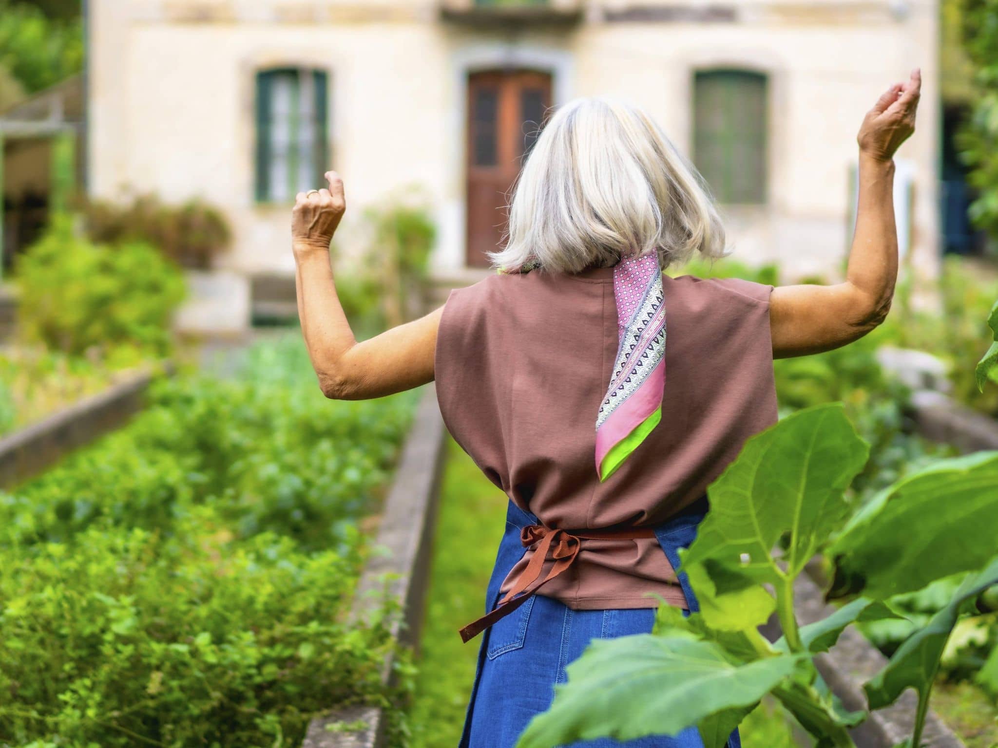 Fitte, ältere Frau von hinten im Garten mit angehobenen Armen.