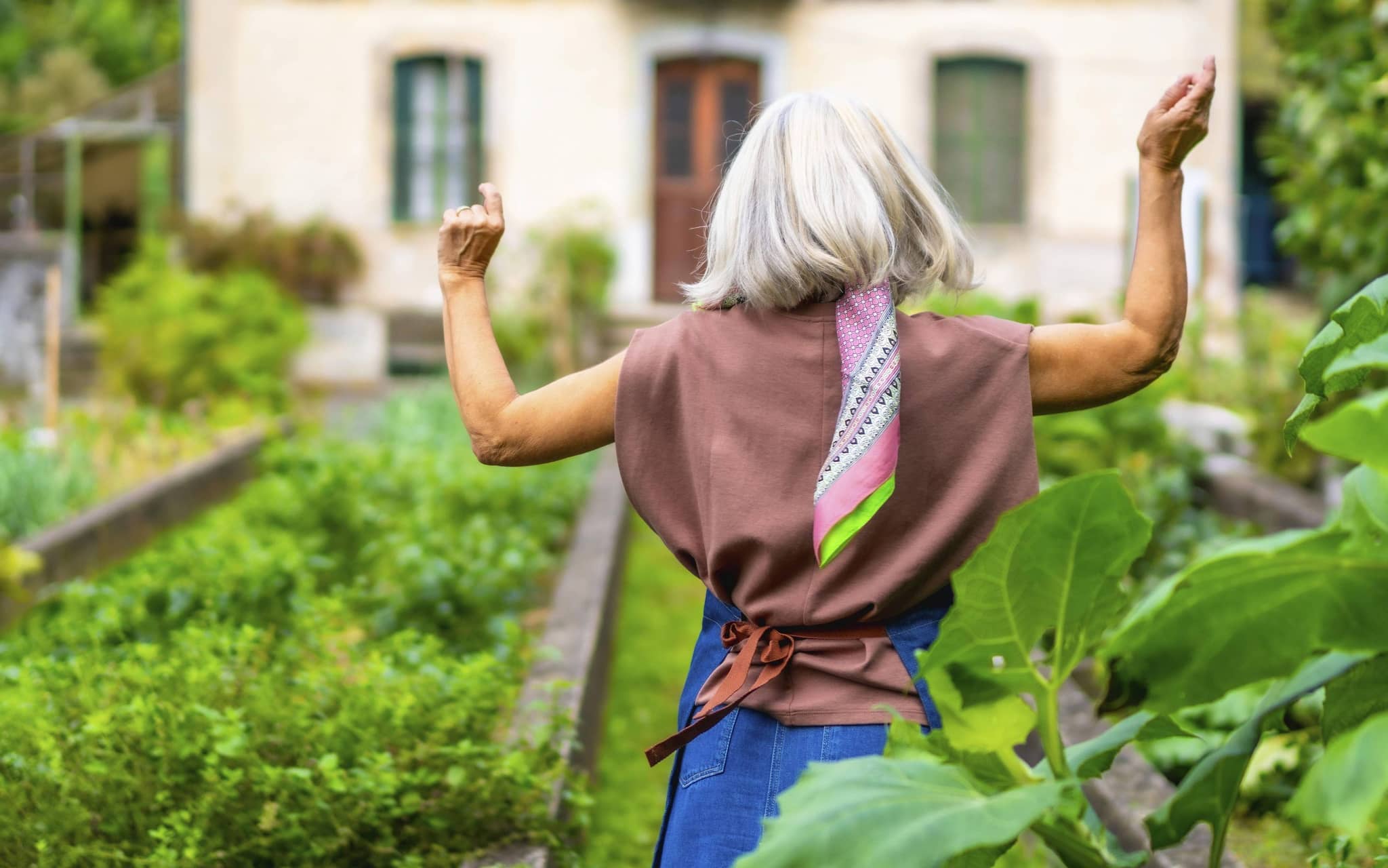 Fitte, ältere Frau von hinten im Garten mit angehobenen Armen.
