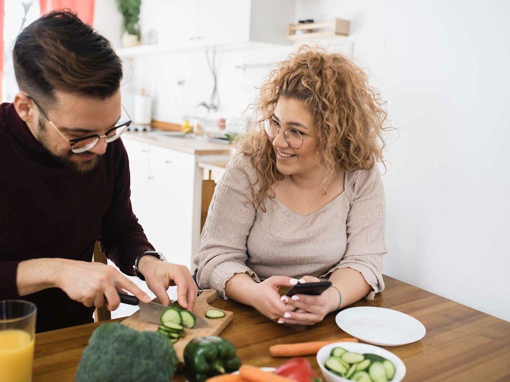 Diabetes-Risiko-Test: Ein junger Mann schneidet Gemüse. Neben ihm sitzt eine junge Frau mit Handy in der Hand.