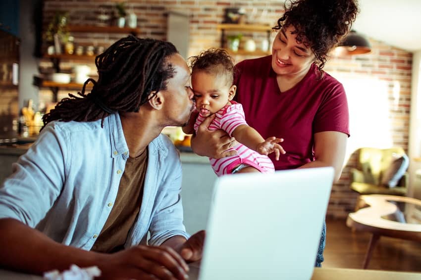 Homeoffice: Vater sitzt arbeitend vor seinem Notebook und küsst sein kleines Kind, das auf dem Arm der Mutter ist.