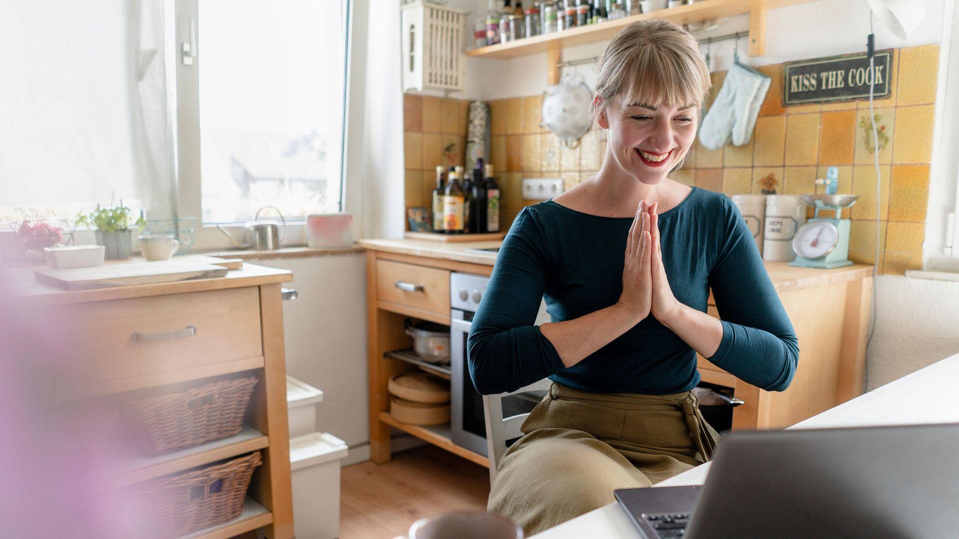 Online-Coaching: Eine Frau sitz in ihrer Küche und lächelt in die Kamera ihres Laptops.