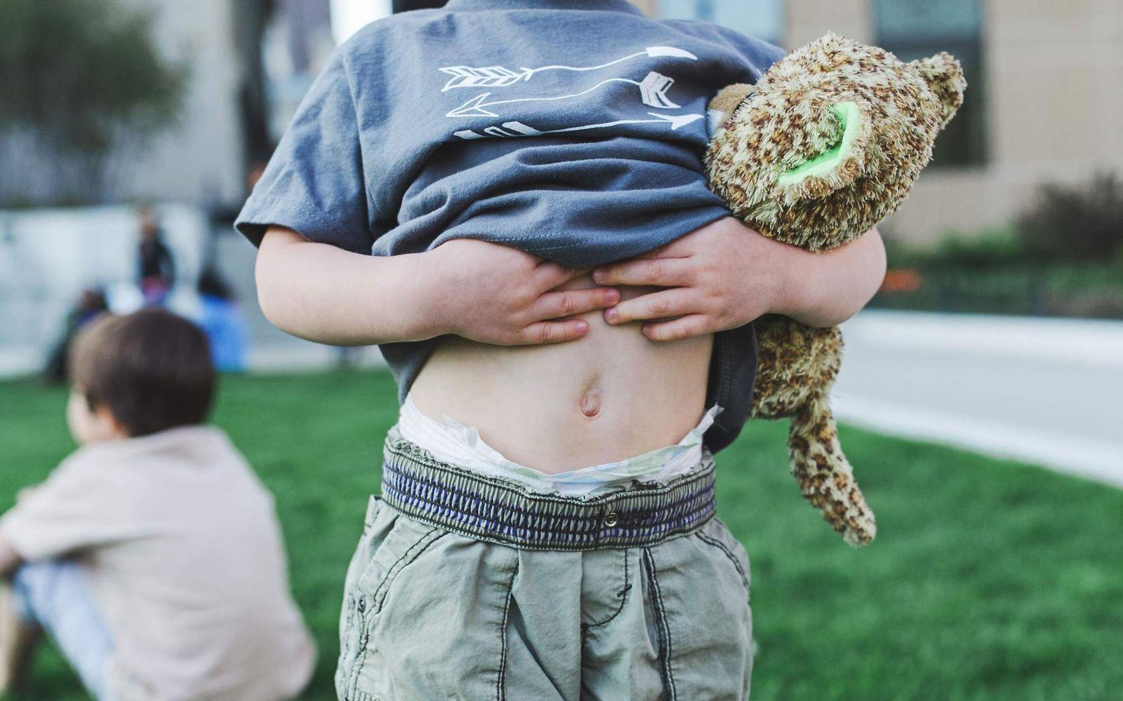 Verstopfung: Kind mit Kuscheltier im Arm zieht das T-Shirt hoch und zeigt seinen nackten Bauch.