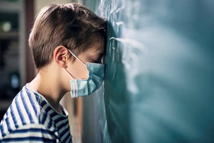 Junge mit Mundschutz steht in der Schule mit geschlossenen Augen vor der Tafel und lehnt den Kopf dagegen.