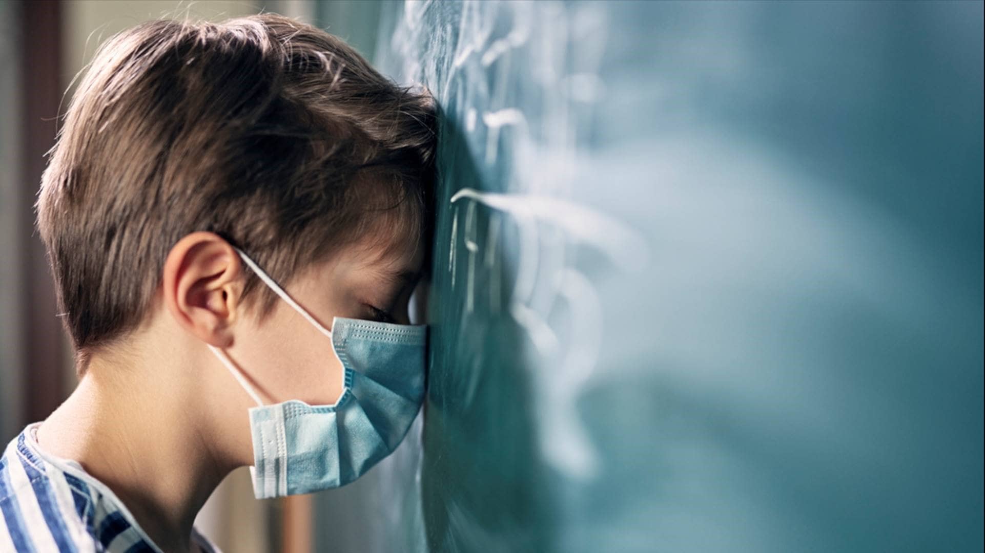 Präventionsradar: Erschöpft wirkendes Kind trägt eine medizinische Maske und stützt seinen Kopf gegen eine Schultafel.