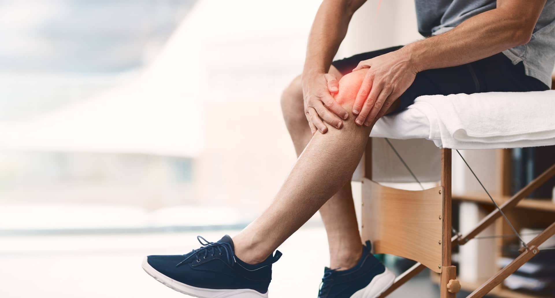 Versorgungsreport Knieschmerz: Mann sitzt auf Behandlungsliege und hält sich das schmerzende Knie.