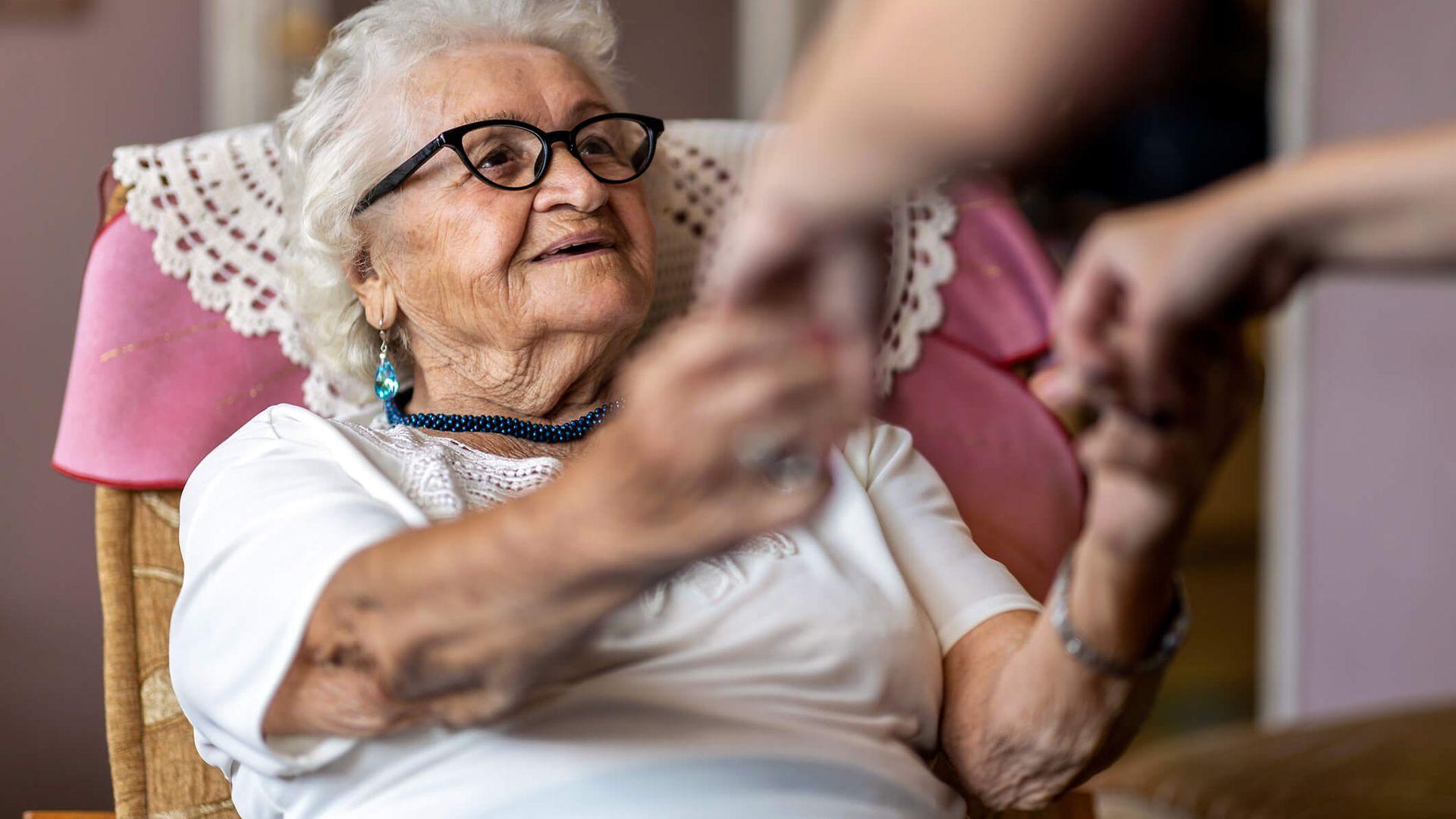 Pflegecoach: Alte Frau sitzt lächelnd im Sessel und bekommt Hilfe beim Aufstehen