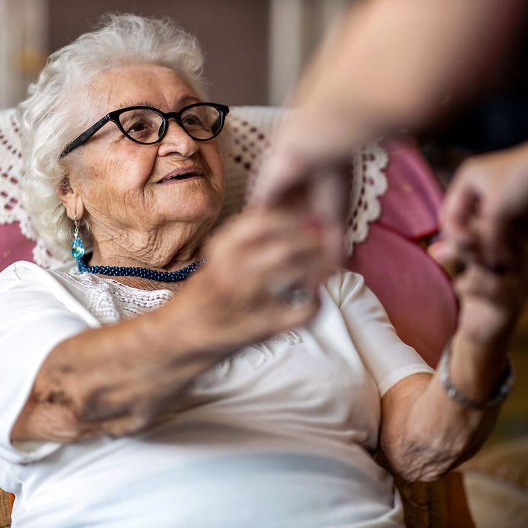 Pflegecoach: Alte Frau sitzt lächelnd im Sessel und bekommt Hilfe beim Aufstehen
