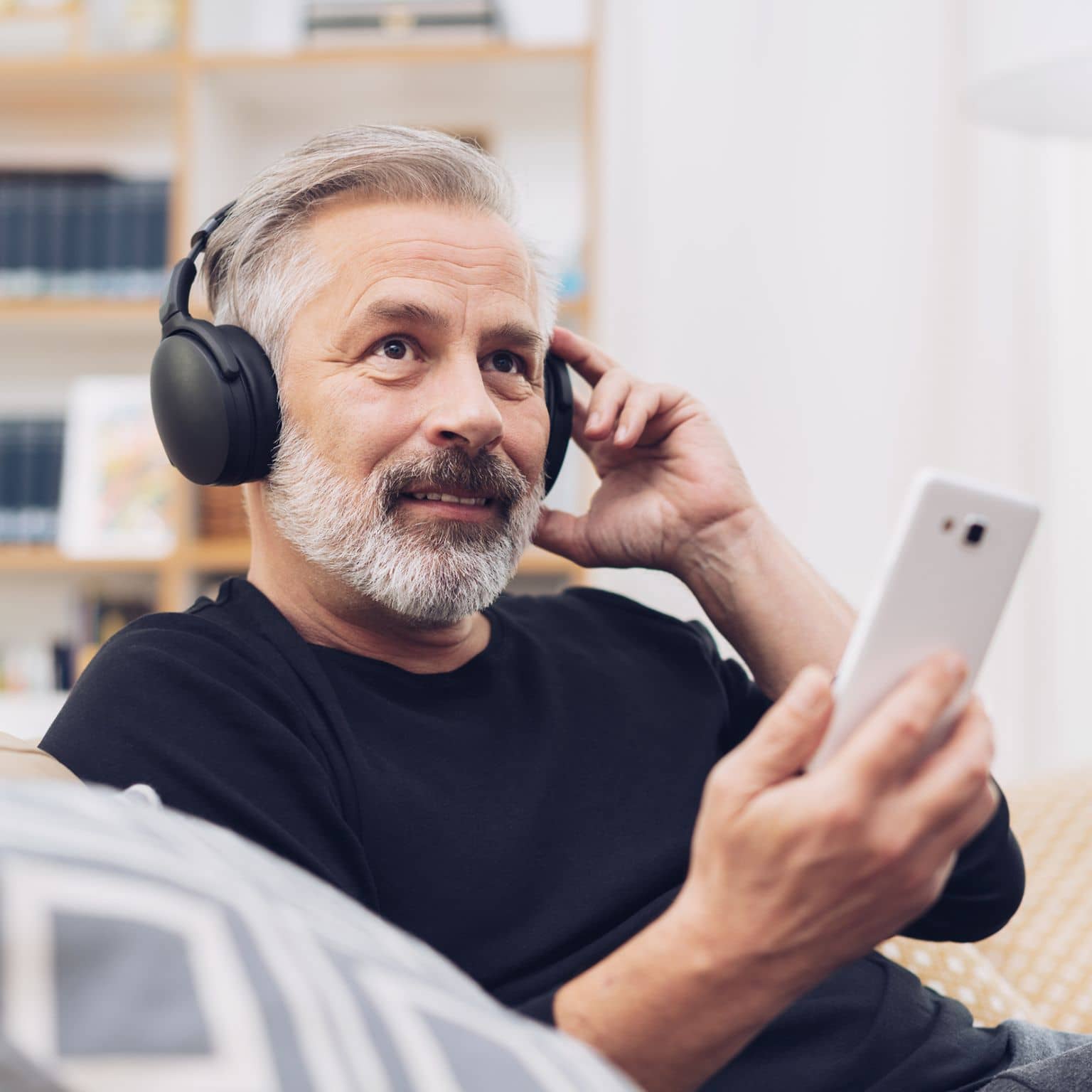 Älterer Mann sitzt auf dem Sofa, hat Kopfhörern auf und sein Handy in der Hand.