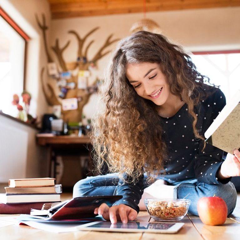 Bild: Jugendliche mit Buch in der Hand sitzt auf dem Boden und bedient ein Tablet.
