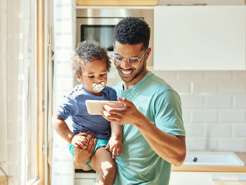 Bild: Mann mit Kind auf dem Arm lächelt ins Smartphone