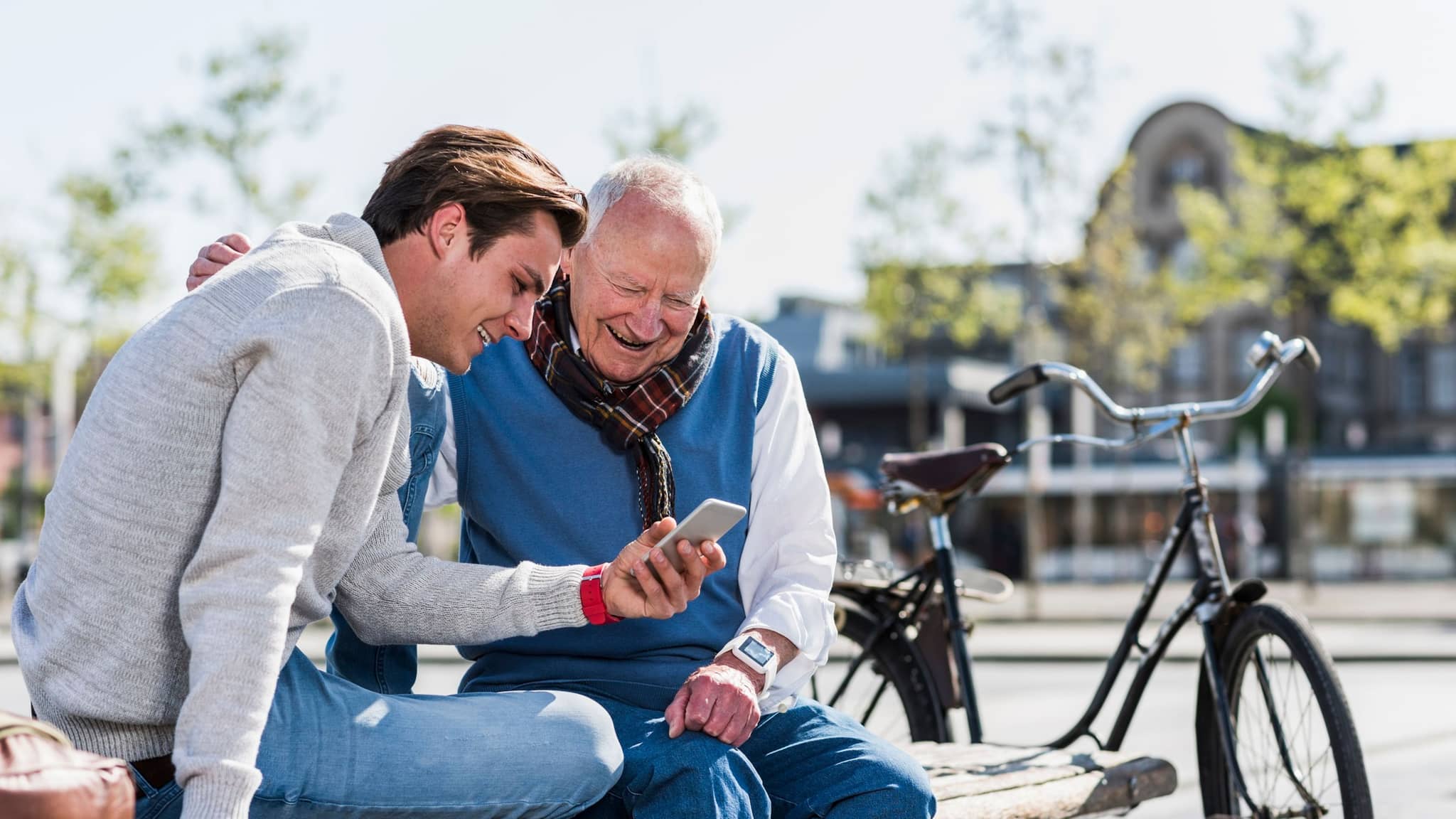 DAK Pflege-App: Enkel sitzt mit Großvater auf einer Parkbank und zeigt lachend auf das Smartphone