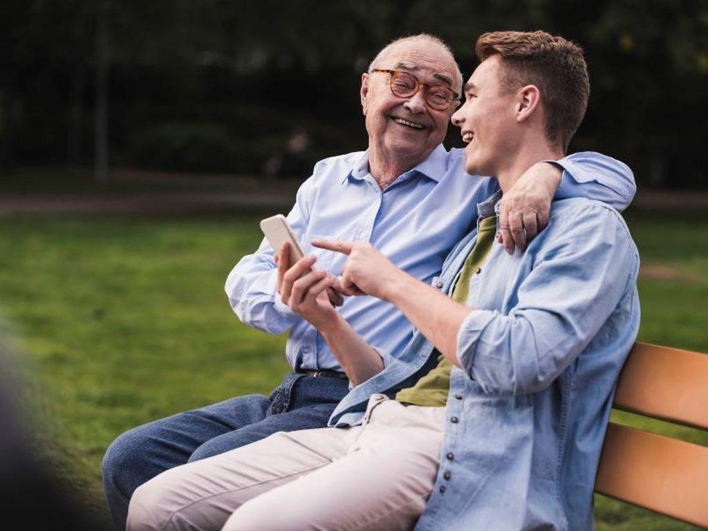 DAK Pflege-App: Enkel sitzt mit Großvater auf einer Parkbank und zeigt lachend auf das Smartphone