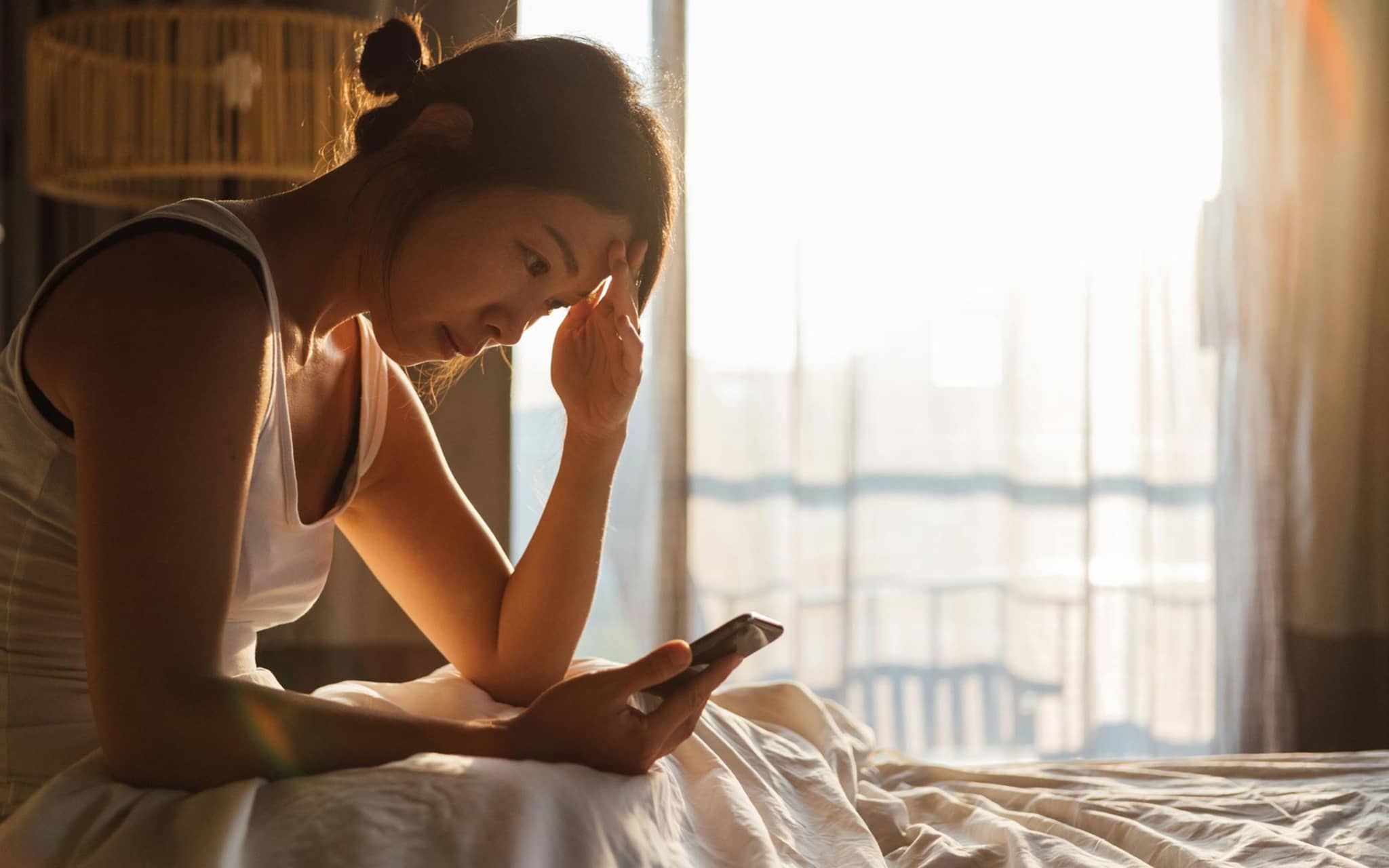 Fimo App bei Long Covid: Junge Frau sitzt erschöpft im Bett und schaut auf ihr Handy.